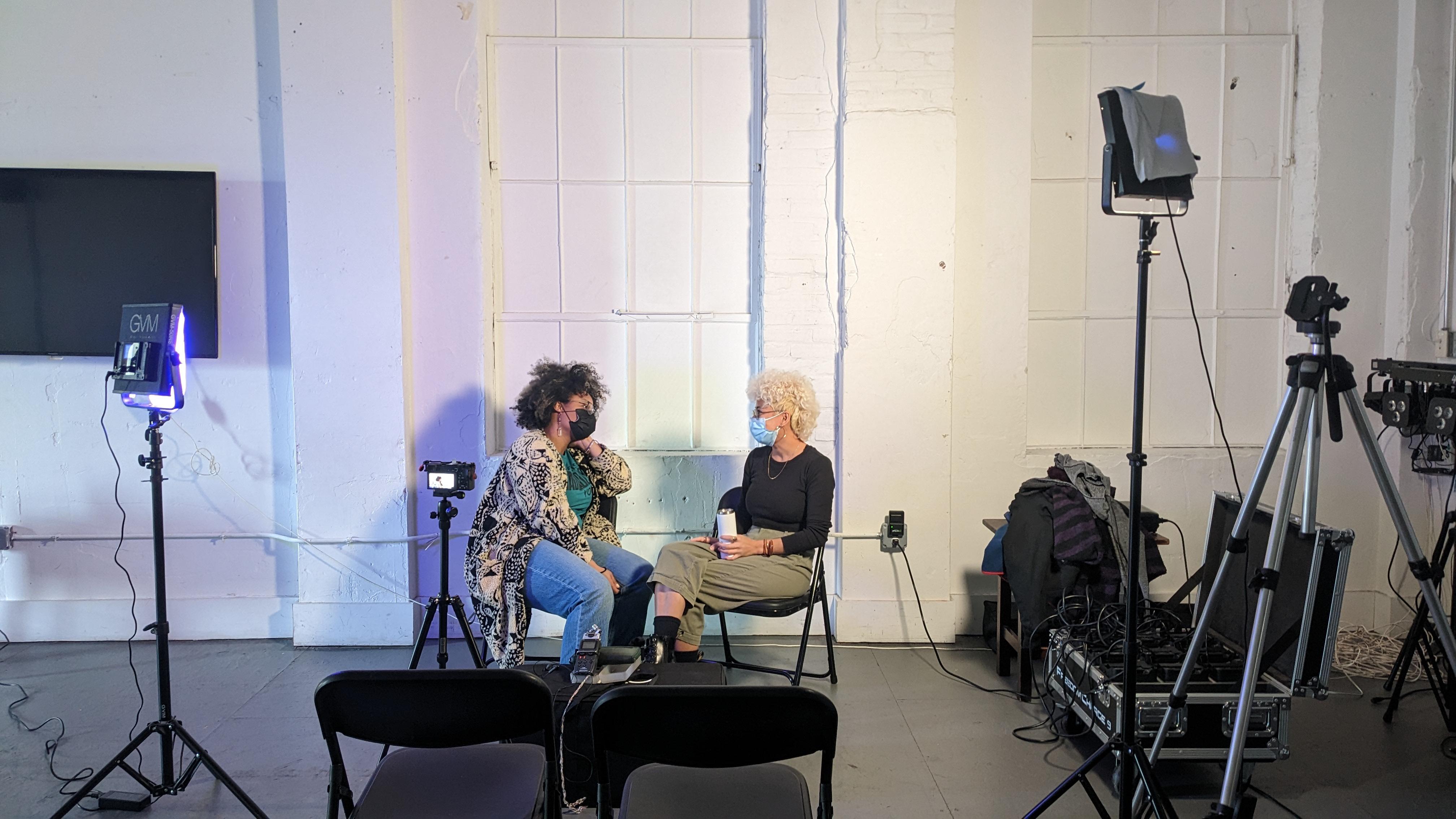 Photo by eo Studios. Two people sit facing each other in a studio surrounded by lighting and video equipment.