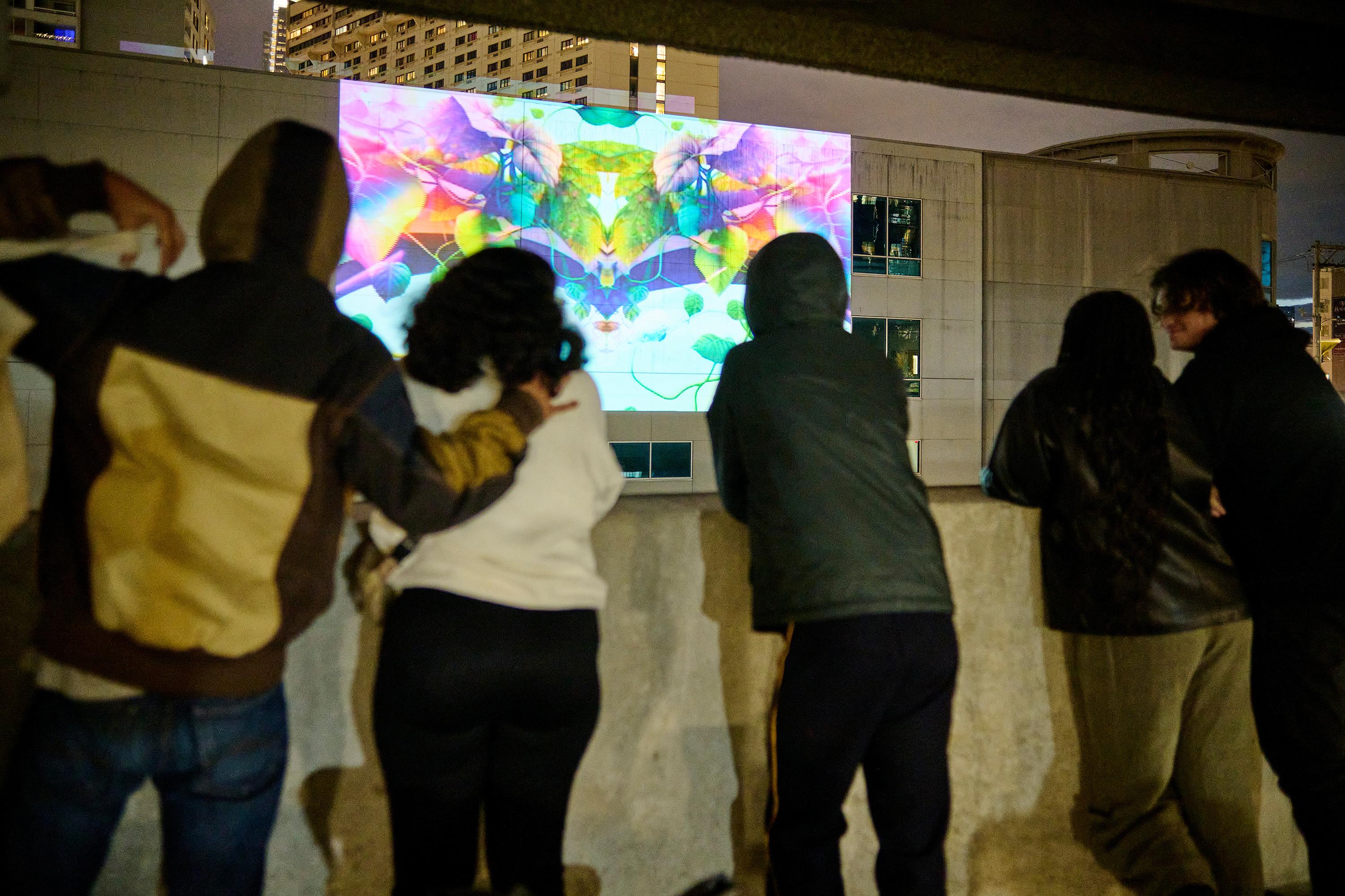 Photo courtesy of Albert Yee. Close up of people at night leaning against the wall looking a colorful projection against a building wall.