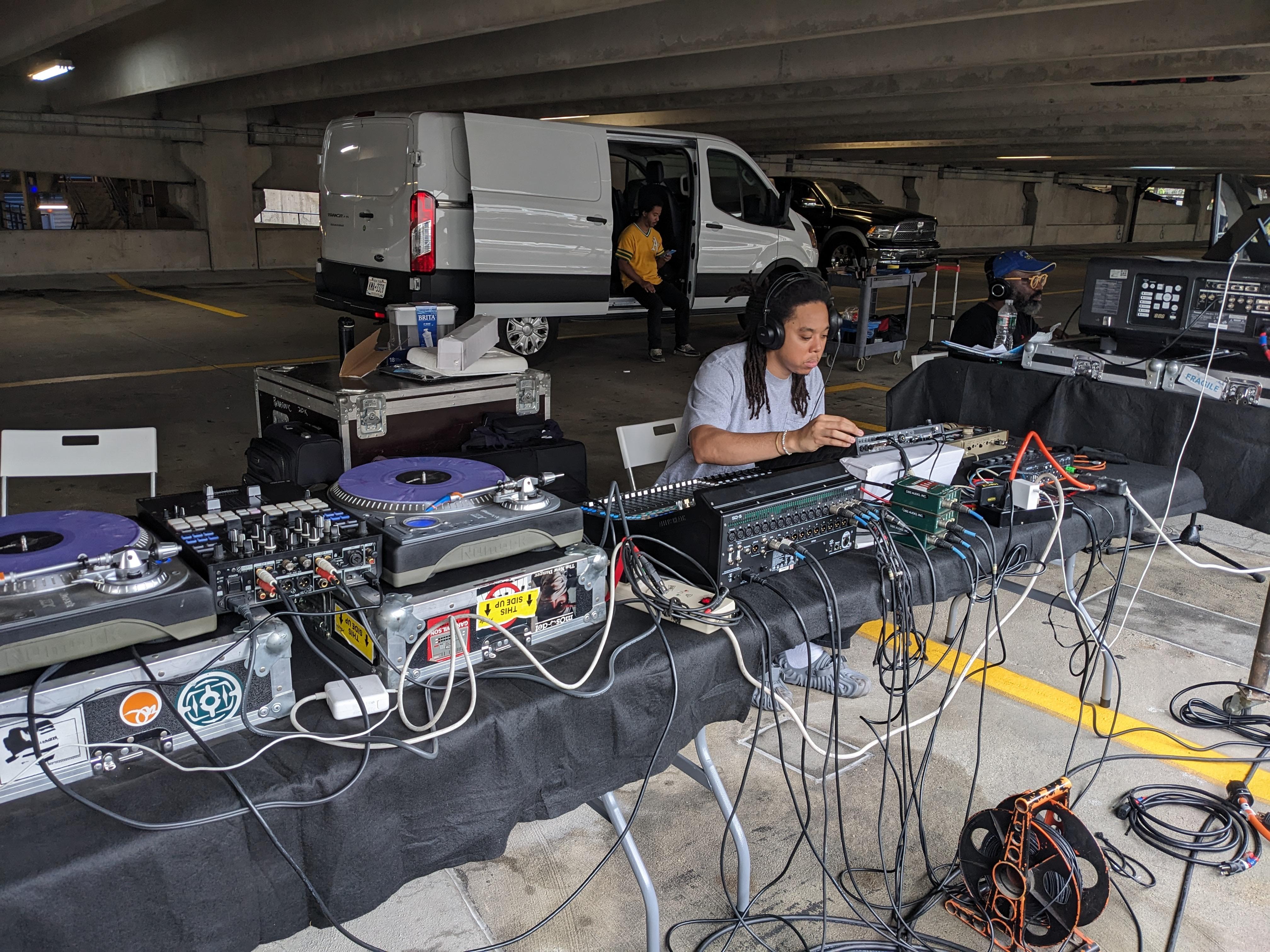 Photo by eo Studios. A person works at a table covered in computers and audio equipment, inside a parking garage.