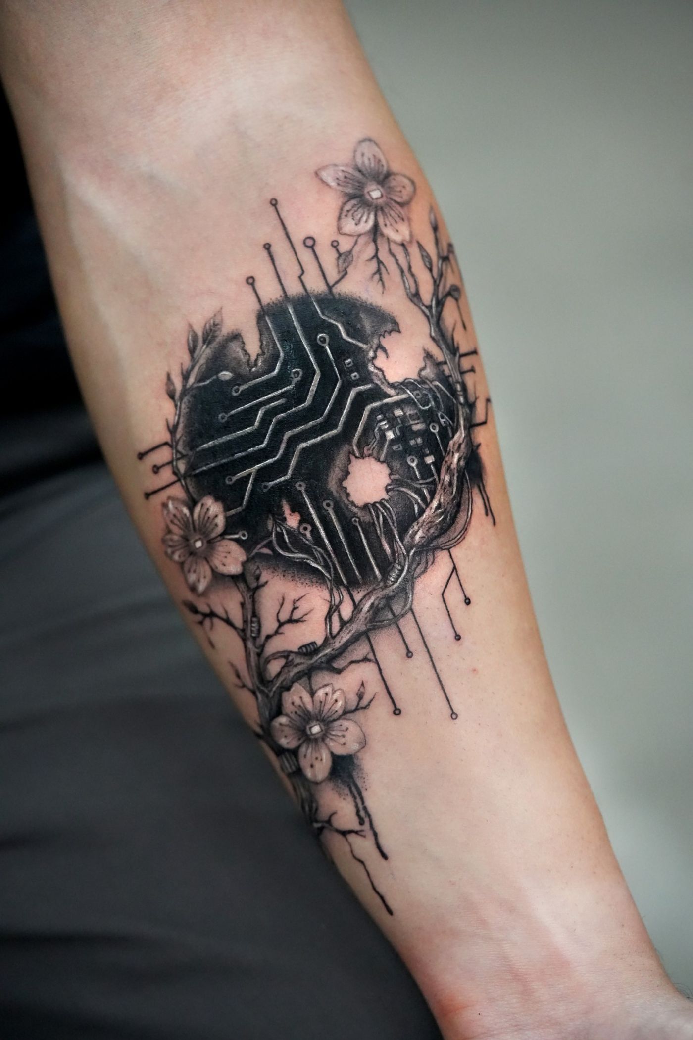 Schwarz-Grau-Tattoo am Unterarm: eine aufgebrochene dunkle Kugel mit Leiterbahnen, umrankt von Kirschblütenzweigen