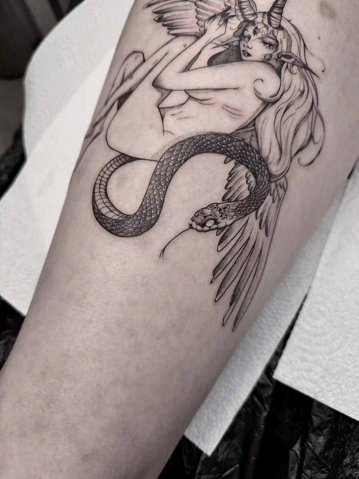 Schwarz-Grau-Tattoo einer gehörnten weiblichen Figur mit langem Haar, elfenartigen Ohren und Flügeln, umschlungen von einer detaillierten Schlange – surreales, fantasy-inspiriertes Fineline-Design am Arm.