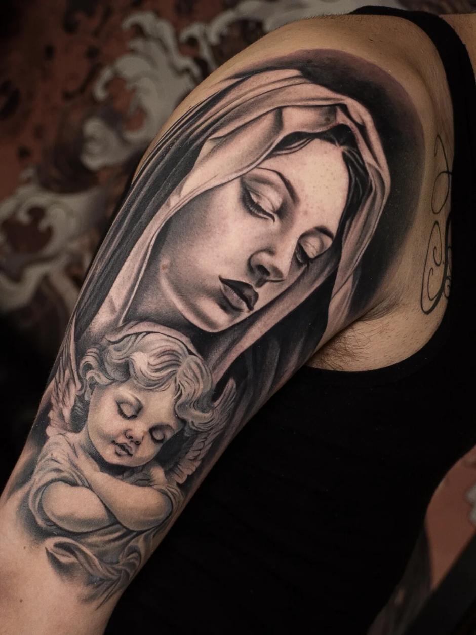Schwarz-Grau-Realismus-Tattoo einer ruhigen Marienfigur, die einen schlafenden Engel umarmt, detailliert mit weichen Schatten und religiöser Symbolik am Oberarm.