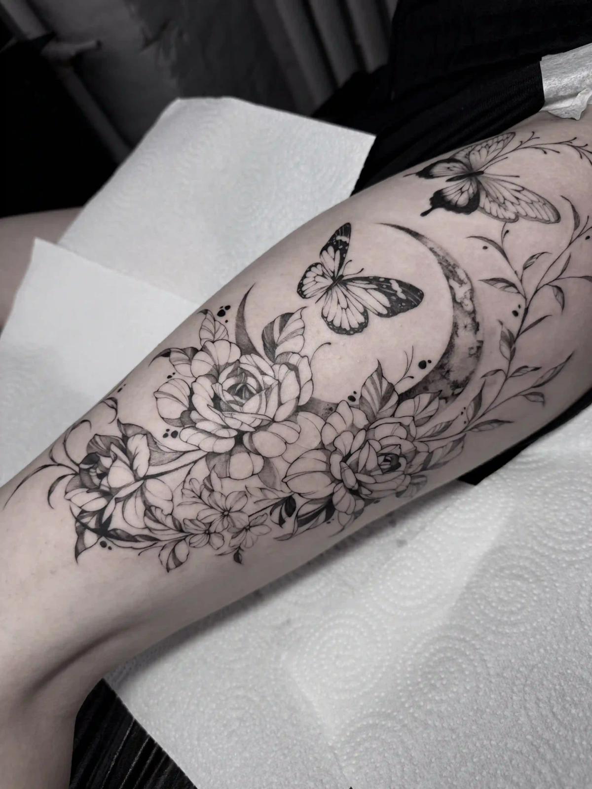 Florales und himmlisches Tattoo mit Rosen, Schmetterlingen und einer Mondsichel, in Schwarz-Grau mit weicher Schattierung und feinen botanischen Fineline-Details am Bein.