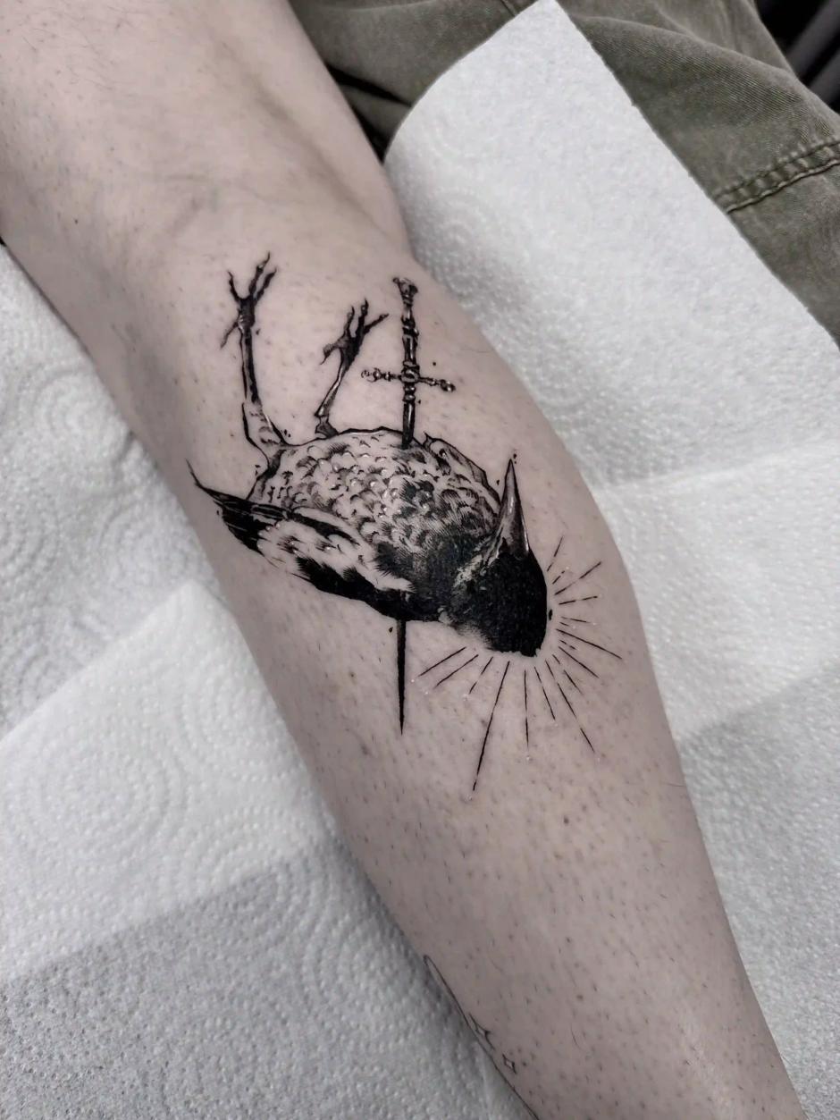 Blackwork-Tattoo eines toten Vogels, der von einem Schwert durchbohrt wird, mit strahlenden Linien als Symbol für Licht oder Einschlag, im ausdrucksstarken Sketch-Stil am Bein.
