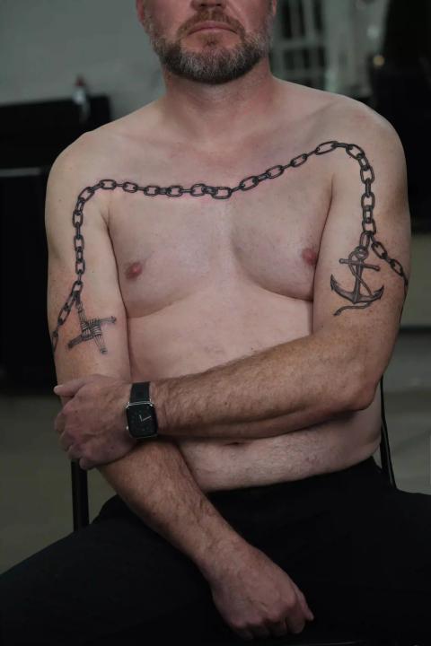 Fineline-Tattoo einer Kette mit Anker und Kreuz über Brust und Armen