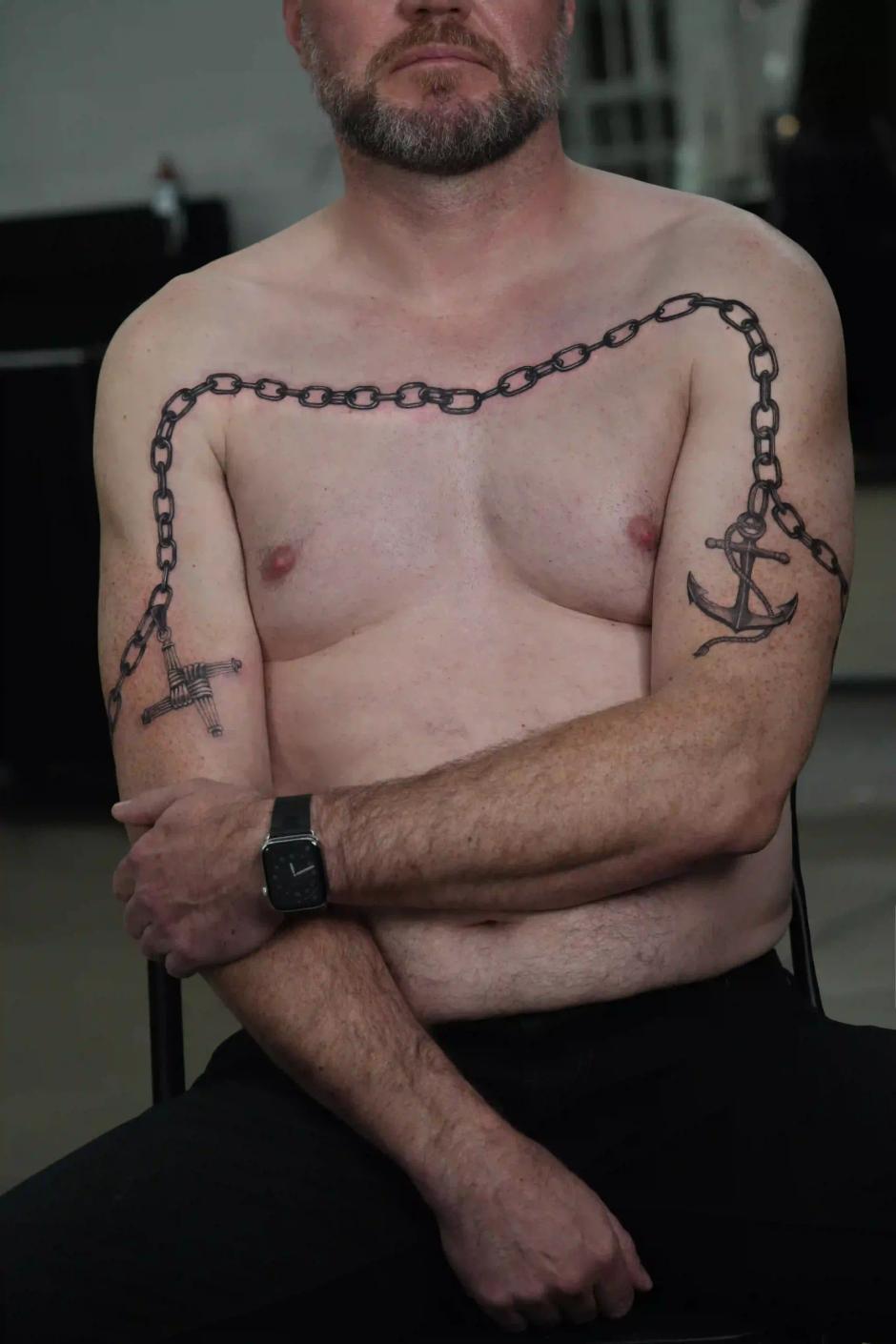 Fineline-Tattoo einer Kette mit Anker und Kreuz über Brust und Armen