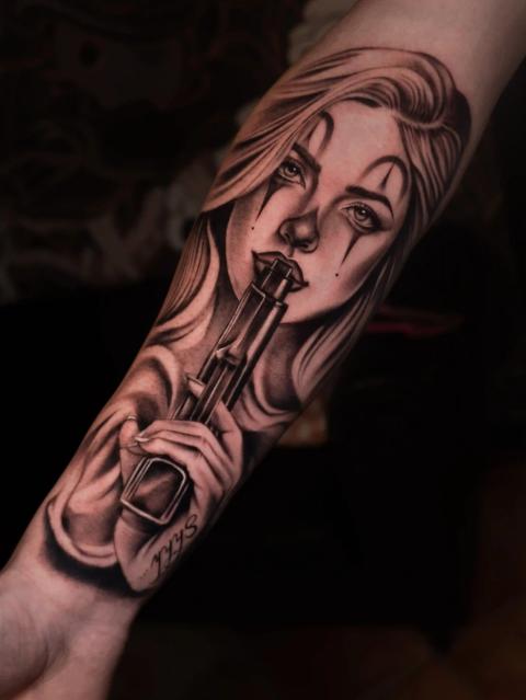 Schwarz-Grau-Realismus-Tattoo im Chicano-Stil eines Frauenporträts mit Clown-Make-up, das eine Waffe an die Lippen hält, mit dramatischem Licht und weicher Schattierung am Unterarm.