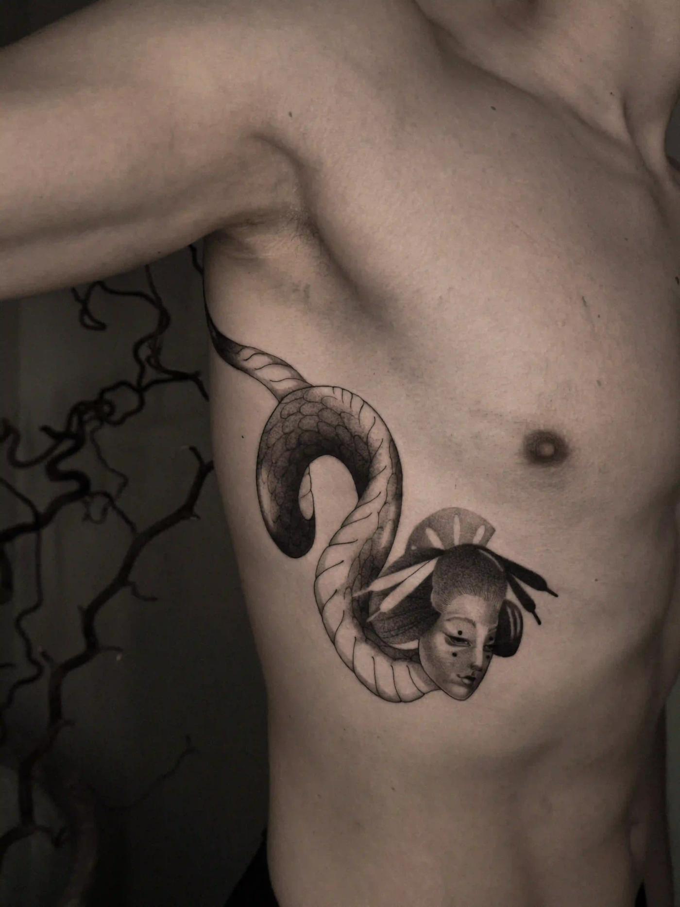 Illustratives Tattoo einer sich windenden Schlange, die aus einer japanisch inspirierten Maske hervortritt, mit Schlangenschuppen und stilisierter Porträt-Schattierung am Rippenbogen.