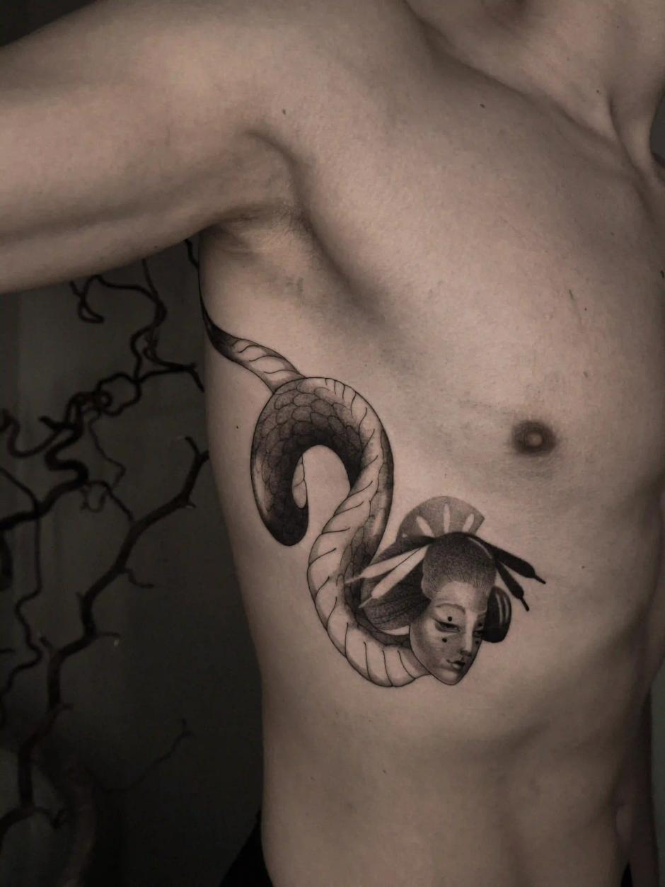 Illustratives Tattoo einer sich windenden Schlange, die aus einer japanisch inspirierten Maske hervortritt, mit Schlangenschuppen und stilisierter Porträt-Schattierung am Rippenbogen.
