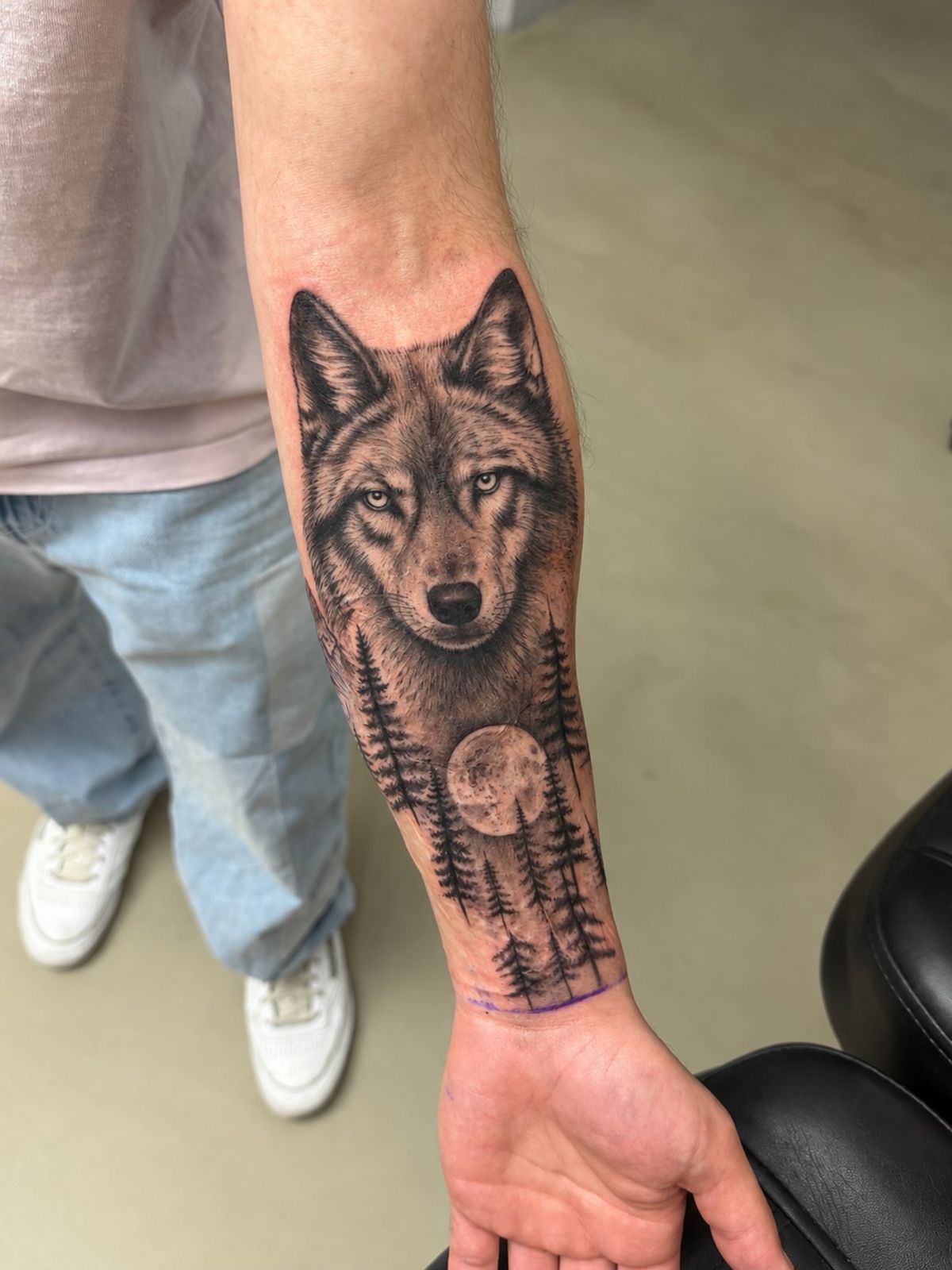 Scar cover-up on the forearm: a realistic black-and-grey wolf tattoo with a full moon and pine forest, tattooed over extensive scarring
