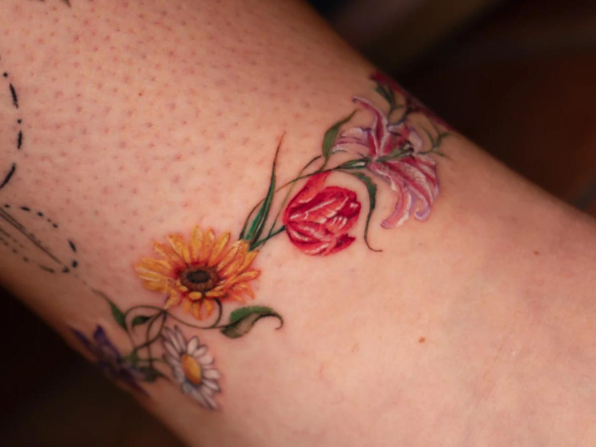Farbenfrohes Mikrorealismus-Blumen-Tattoo mit Gänseblümchen, Tulpen, Lilien und Sonnenblumen im Fineline-Aquarellstil am Knöchel.