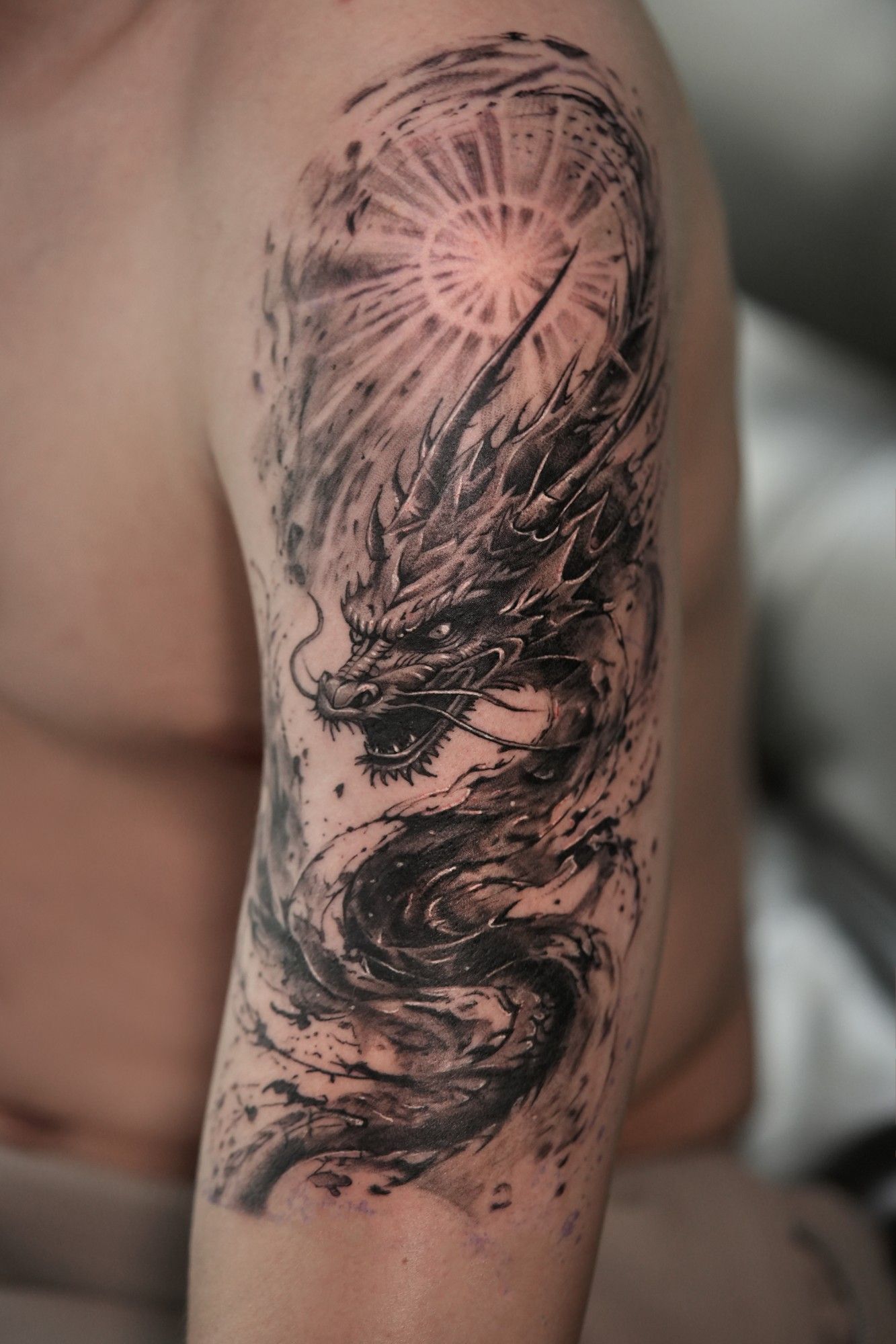 Schwarz-Grau-Tattoo am Oberarm: japanischer Drache in wirbelnder Tusche unter einer strahlenden Sonne