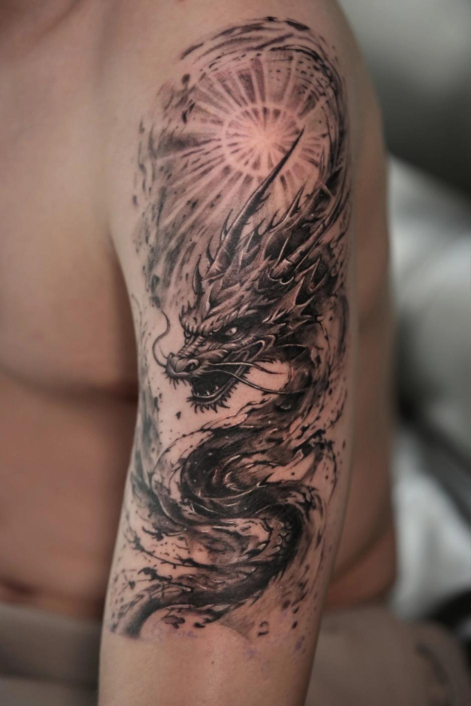 Black and grey upper-arm tattoo: a Japanese dragon in swirling ink beneath a radiant sun