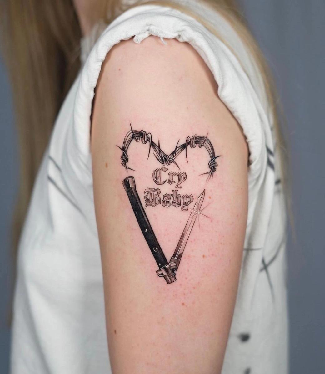 Blackwork-Tattoo zweier Springmesser, die ein V bilden, unter herzförmigem Stacheldraht, mit dem Schriftzug "Cry Baby" in gotischer Schrift am Oberarm.