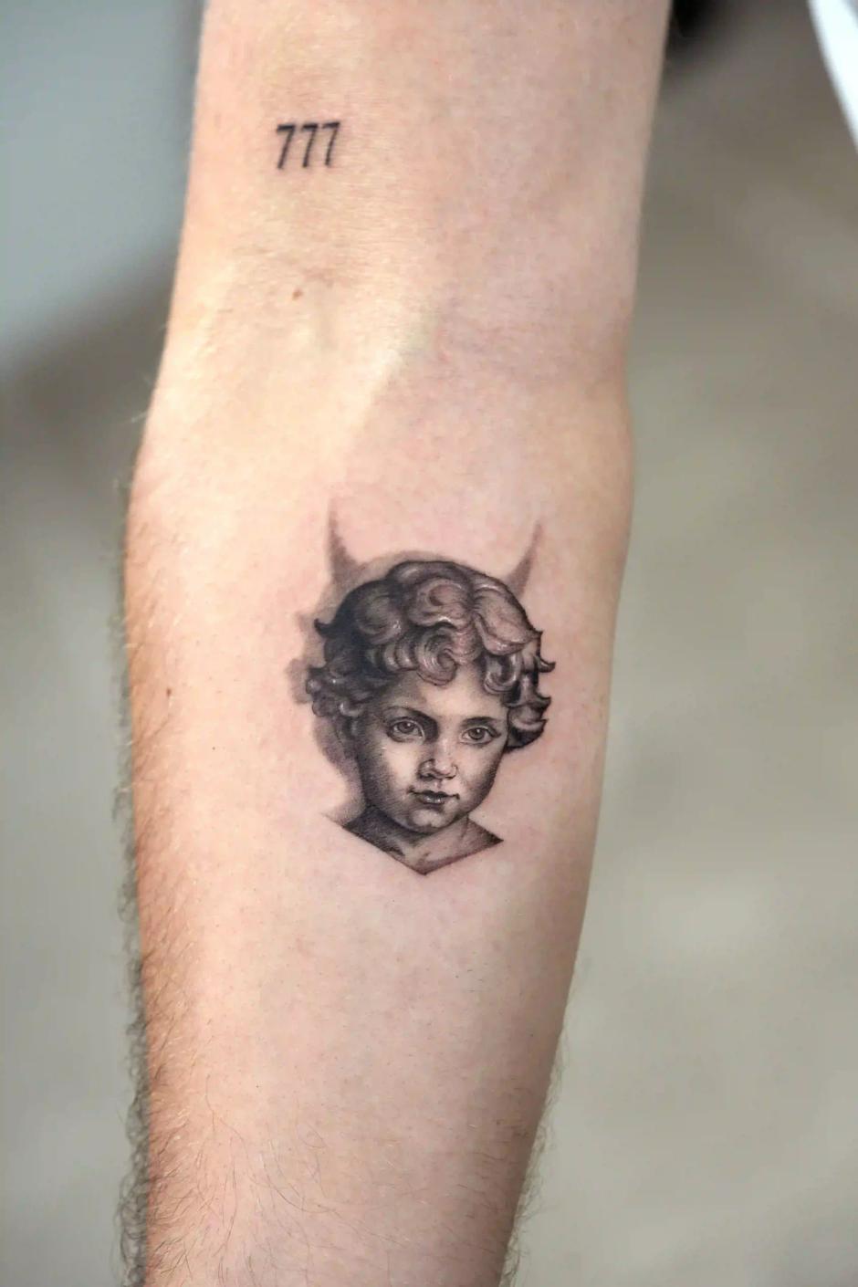 Schwarz-Grau-Mikrorealismus-Tattoo eines Puttenkopfes mit der Zahl '777' am Unterarm