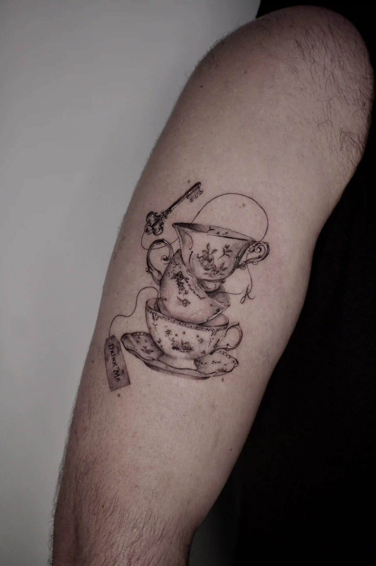 Feines Fineline-Mikrorealismus-Tattoo gestapelter Teetassen mit floralen Motiven, einem Schlüssel und einem Teebeutel-Schild mit der Aufschrift "Drink Me", inspiriert von Alice im Wunderland.