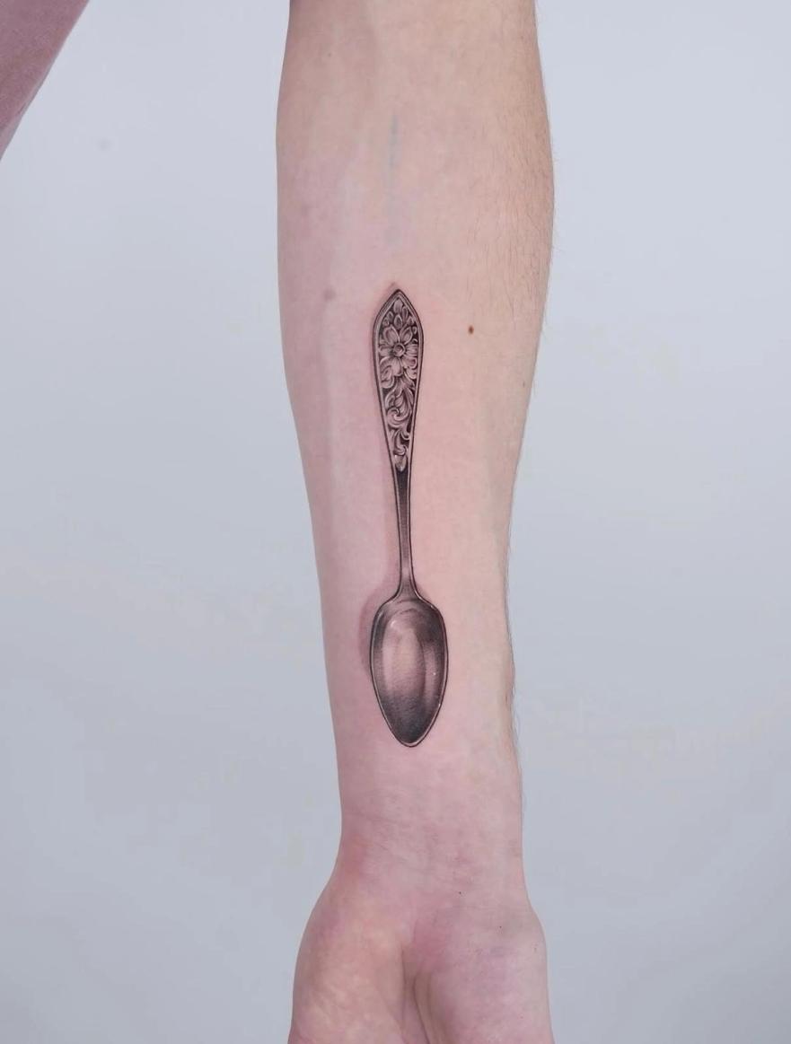 Forearm tattoo: a realistically shaded silver spoon with a floral engraved handle