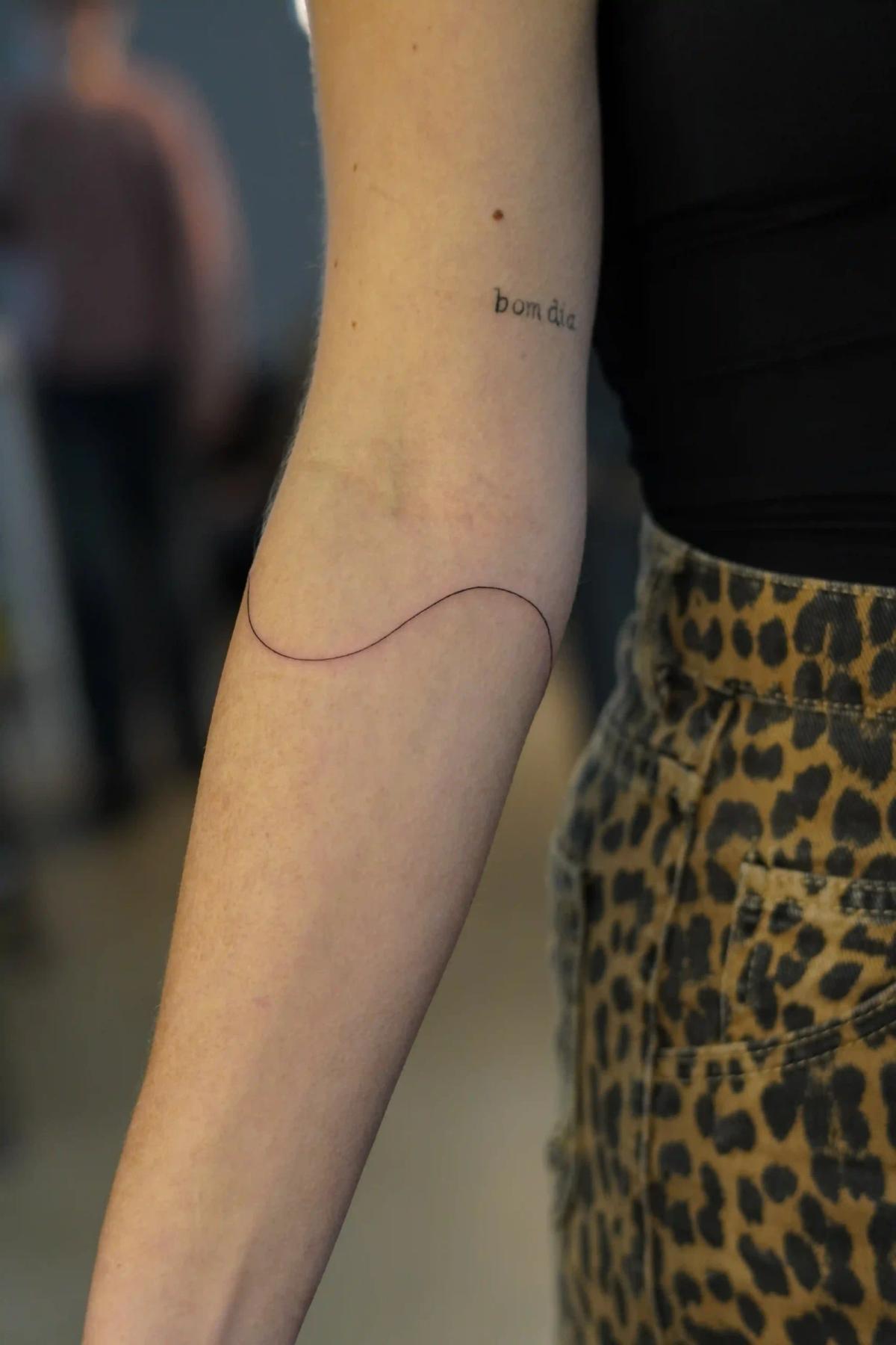 Fineline-Tattoo mit dem Schriftzug 'bom dia' und geschwungener Linie am Unterarm