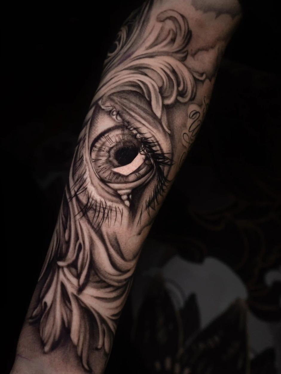Realistisches Schwarz-Grau-Tattoo eines detaillierten menschlichen Auges, umgeben von ornamentalen Barock-Verzierungen, mit Tiefe und Kontrast am Unterarm.