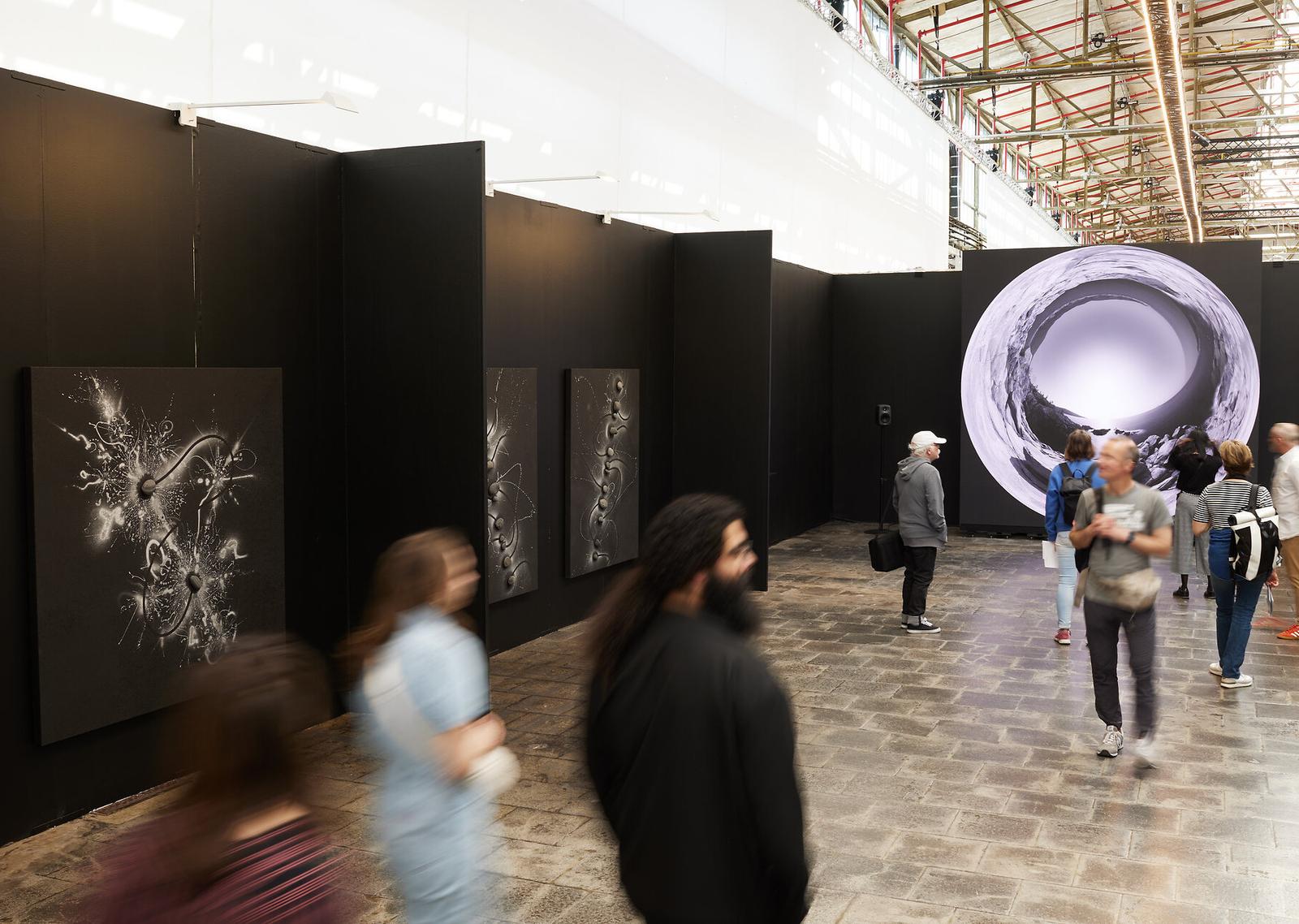 Präsentation des anonymous art project auf der Art Düsseldorf 2025: schwarze Stellwände mit grafischen Arbeiten und großer Videoprojektion.