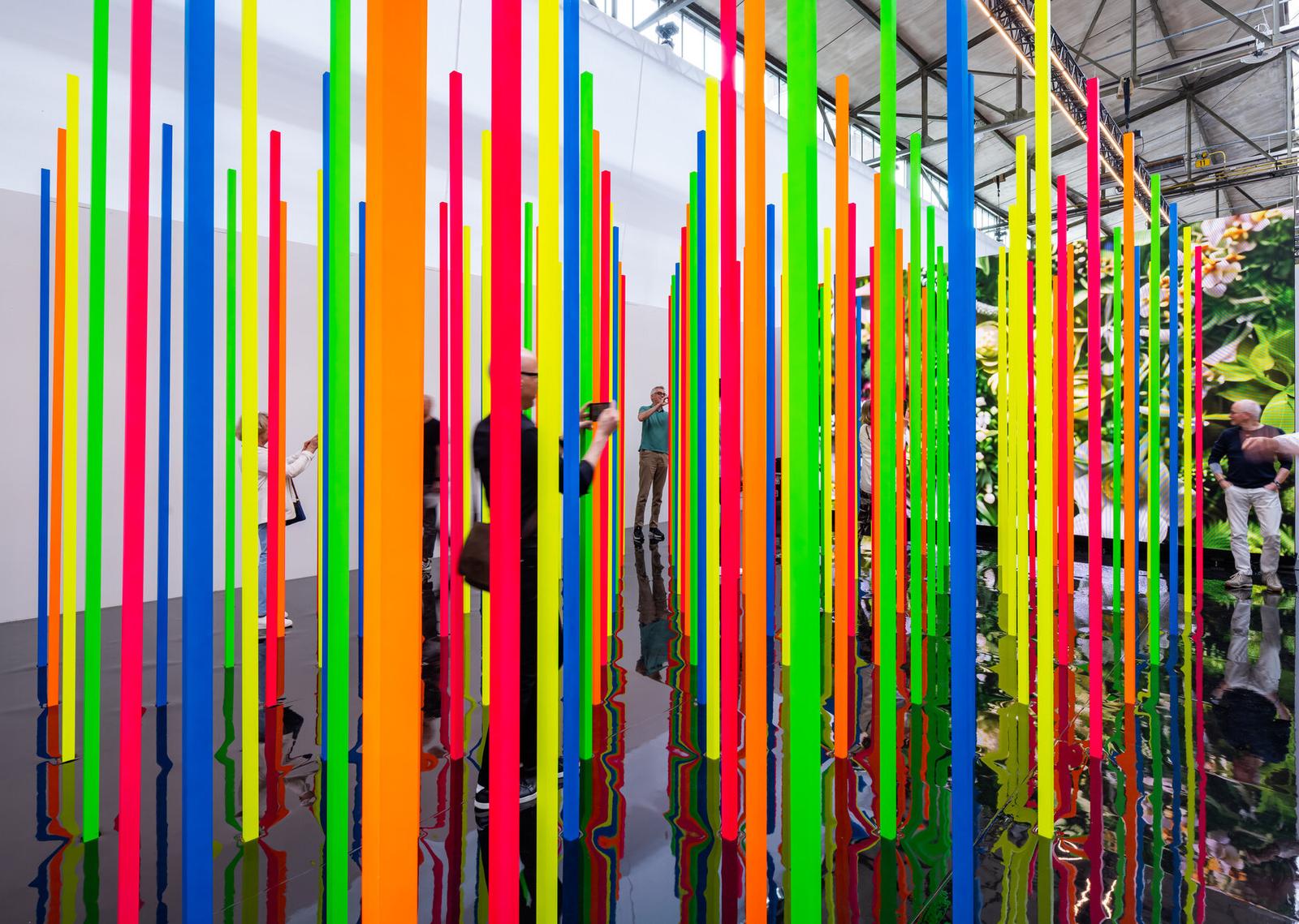 Präsentation des anonymous art project auf der Art Düsseldorf 2024: farbige senkrechte Stäbe auf spiegelndem Boden vor einer Videowand.
