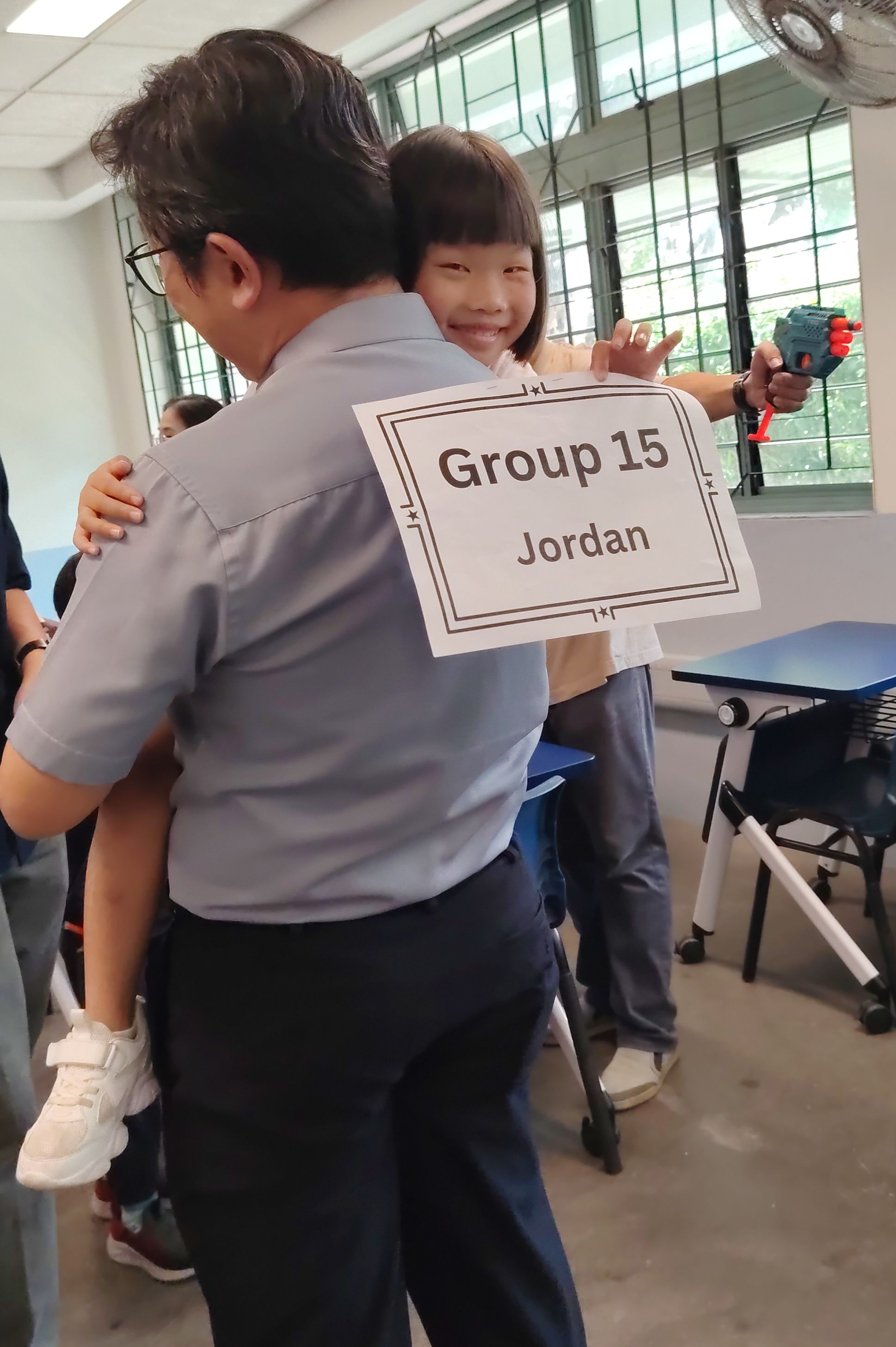Young girl being held up by her father, smiling at the camera while holding out a piece of paper with a group number on it