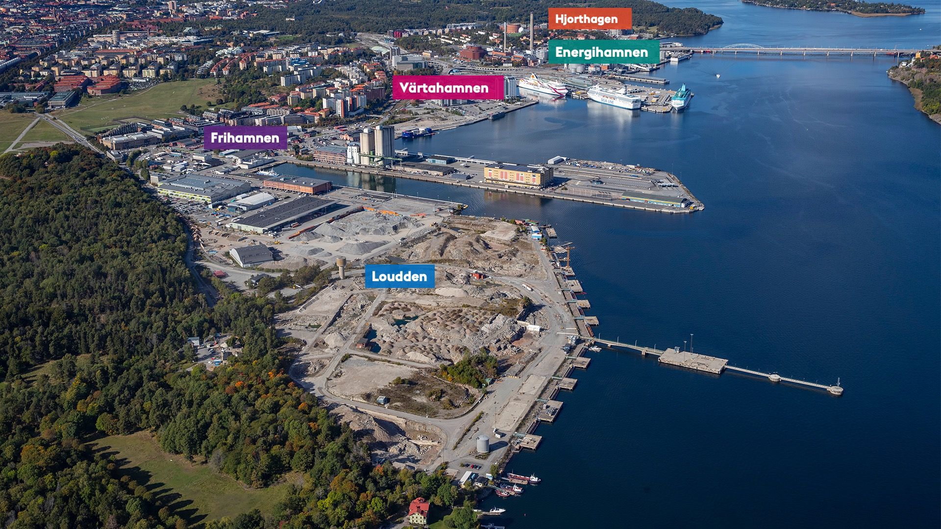 Flygfoto över hamnen i en urban miljö.  Loudden i förgrunden och Frihamnen, Värtahamnen, Energihamnen och Hjorthagen i bakgrunden med en vattenfront som löper längs dessa områden med två fartyg.