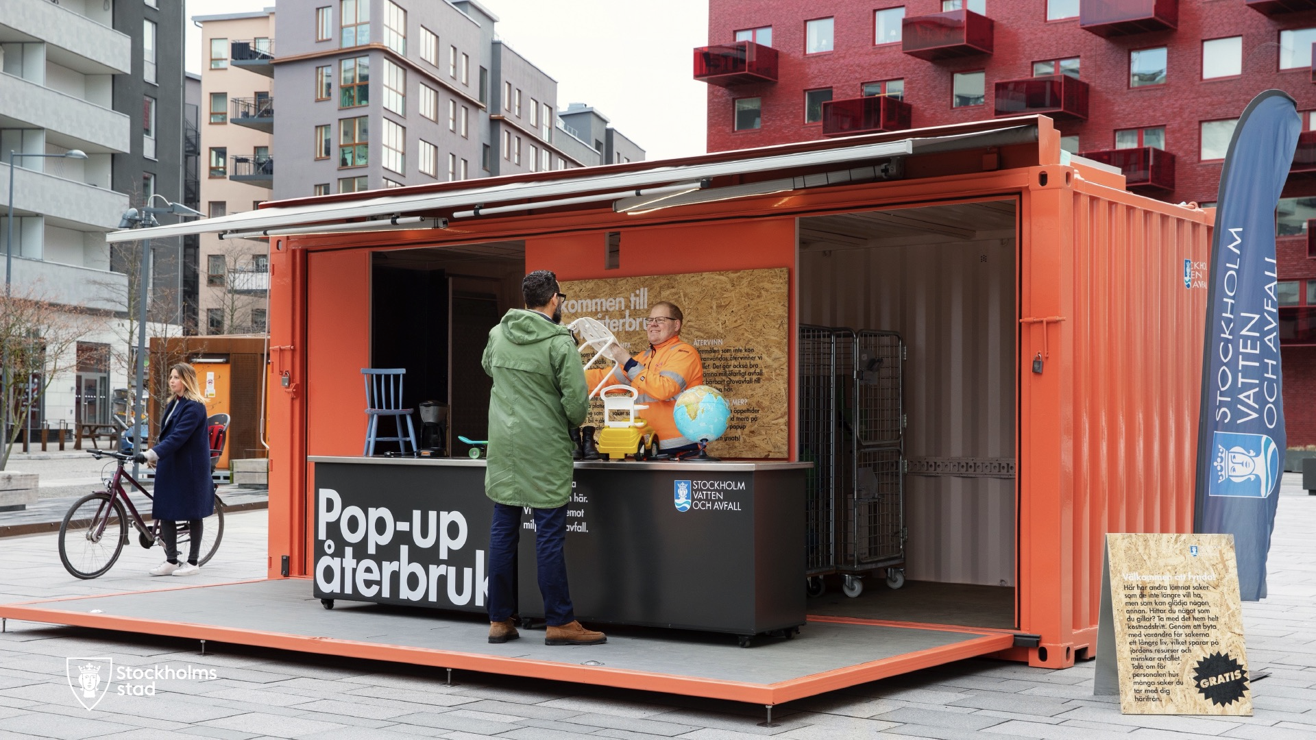 Orange container märkt 'Pop-up återbruk' där en besökare lämnar in föremål till personal i orange väst, med flerbostadshus i bakgrunden.