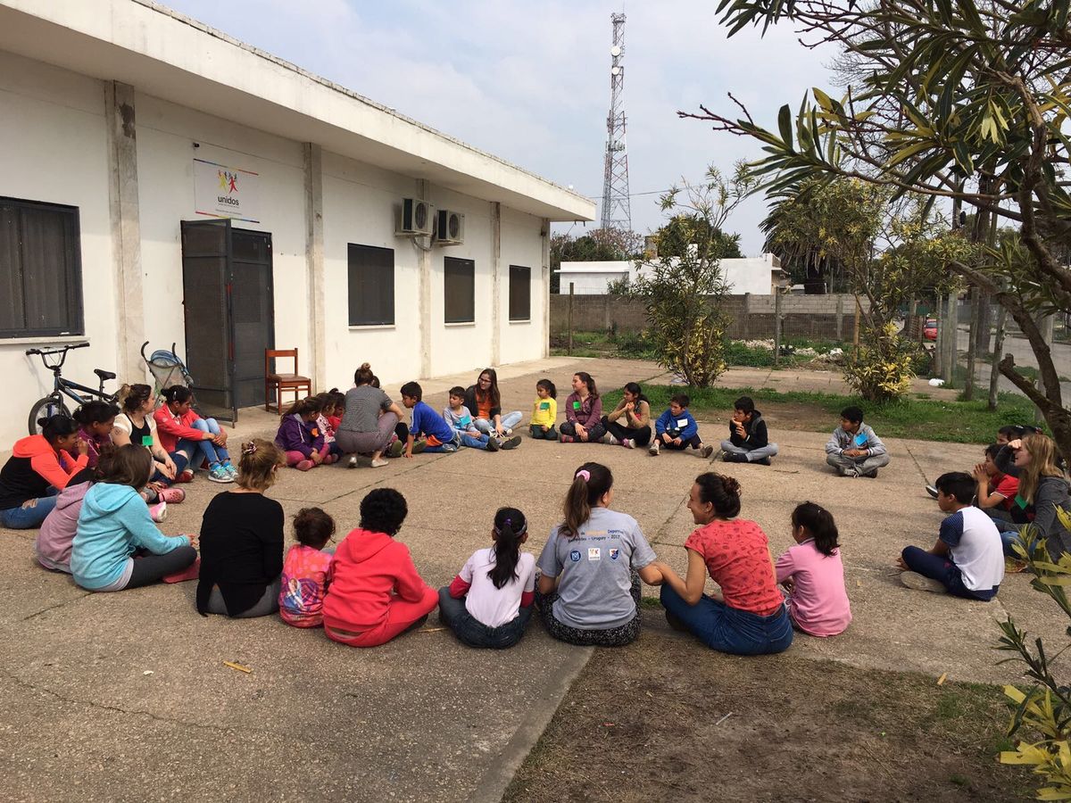 Grupo de niños y adultos sentados en círculo en el patio de un centro comunitario durante una jornada deportiva en Uruguay.