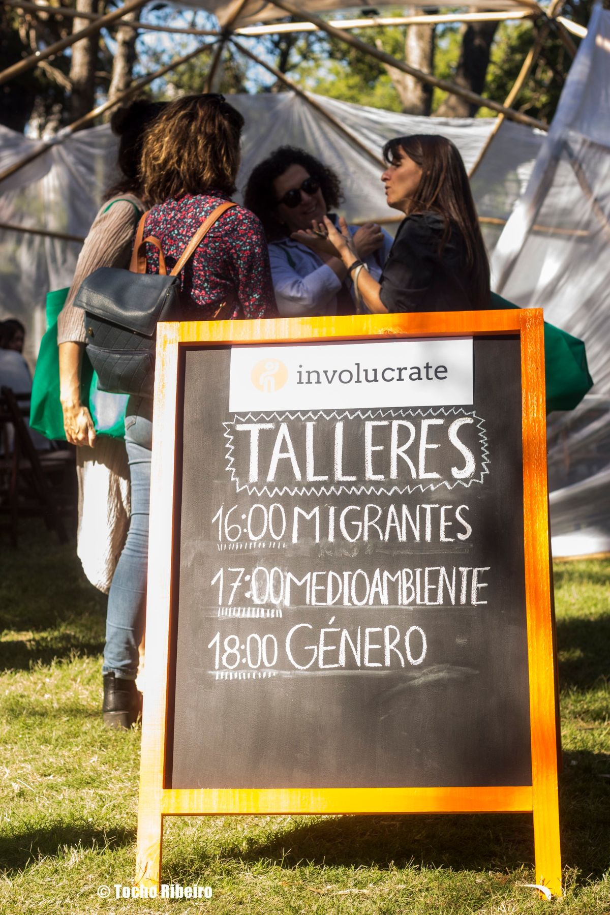 Pizarra con agenda de talleres comunitarios sobre migrantes, medioambiente y género, con voluntarias conversando al aire libre.