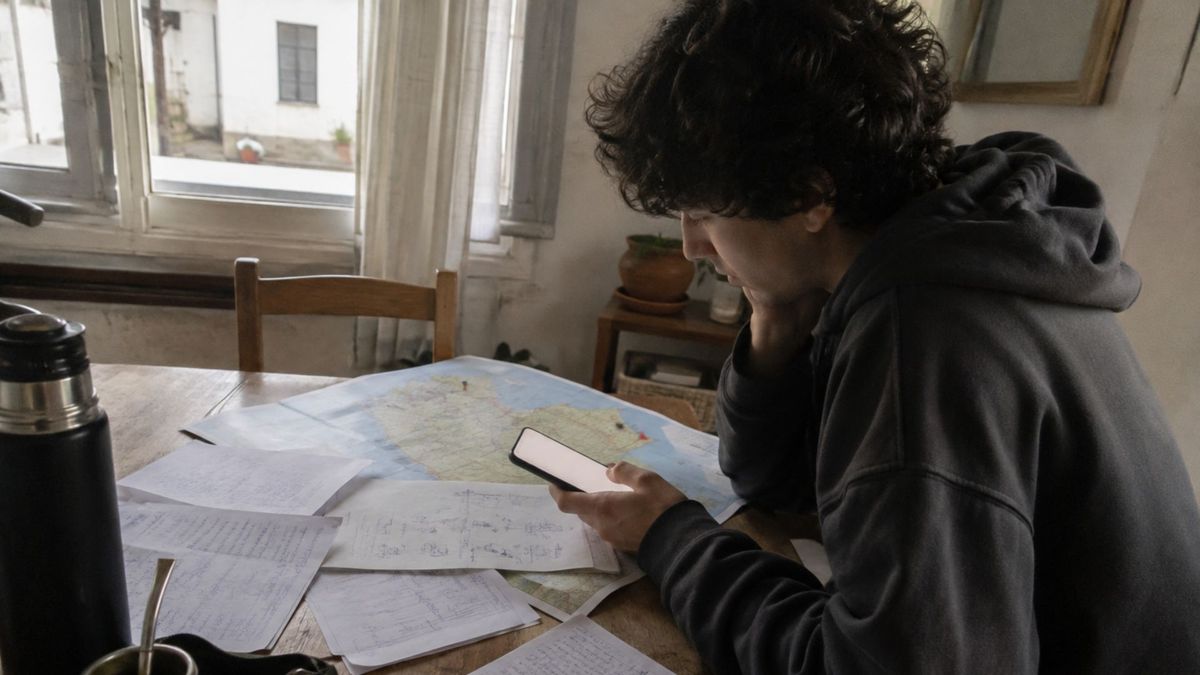 Persona joven sentada en una mesa de madera mira su teléfono mientras revisa notas manuscritas y un mapa impreso de Uruguay con marcas de ubicación, buscando dónde hacer voluntariado.
