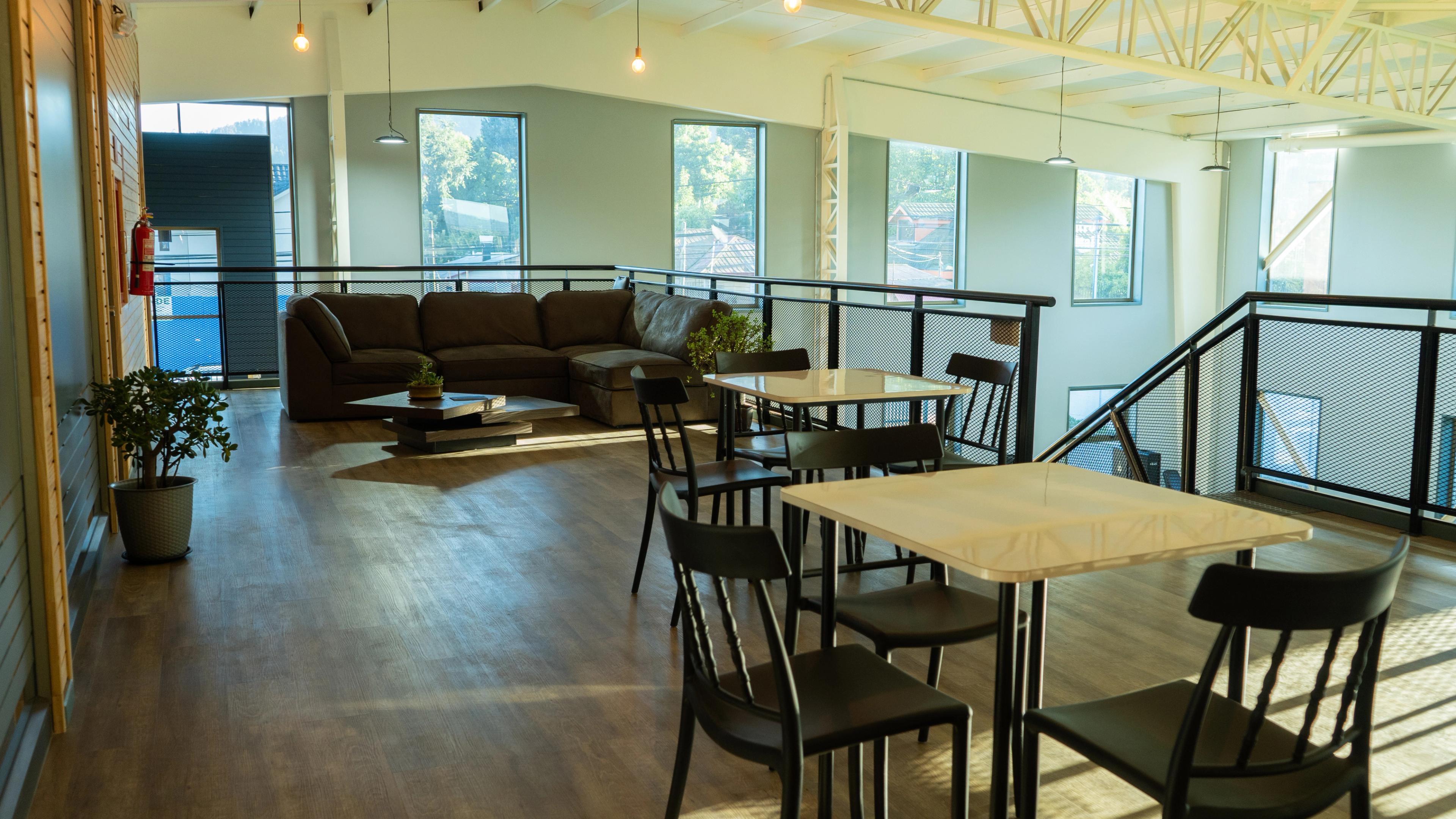 Despacho luminoso con grandes ventanas, suelo de madera y decoración minimalista. Cuenta con mesas y sillas negras, un sofá marrón y plantas en macetas.