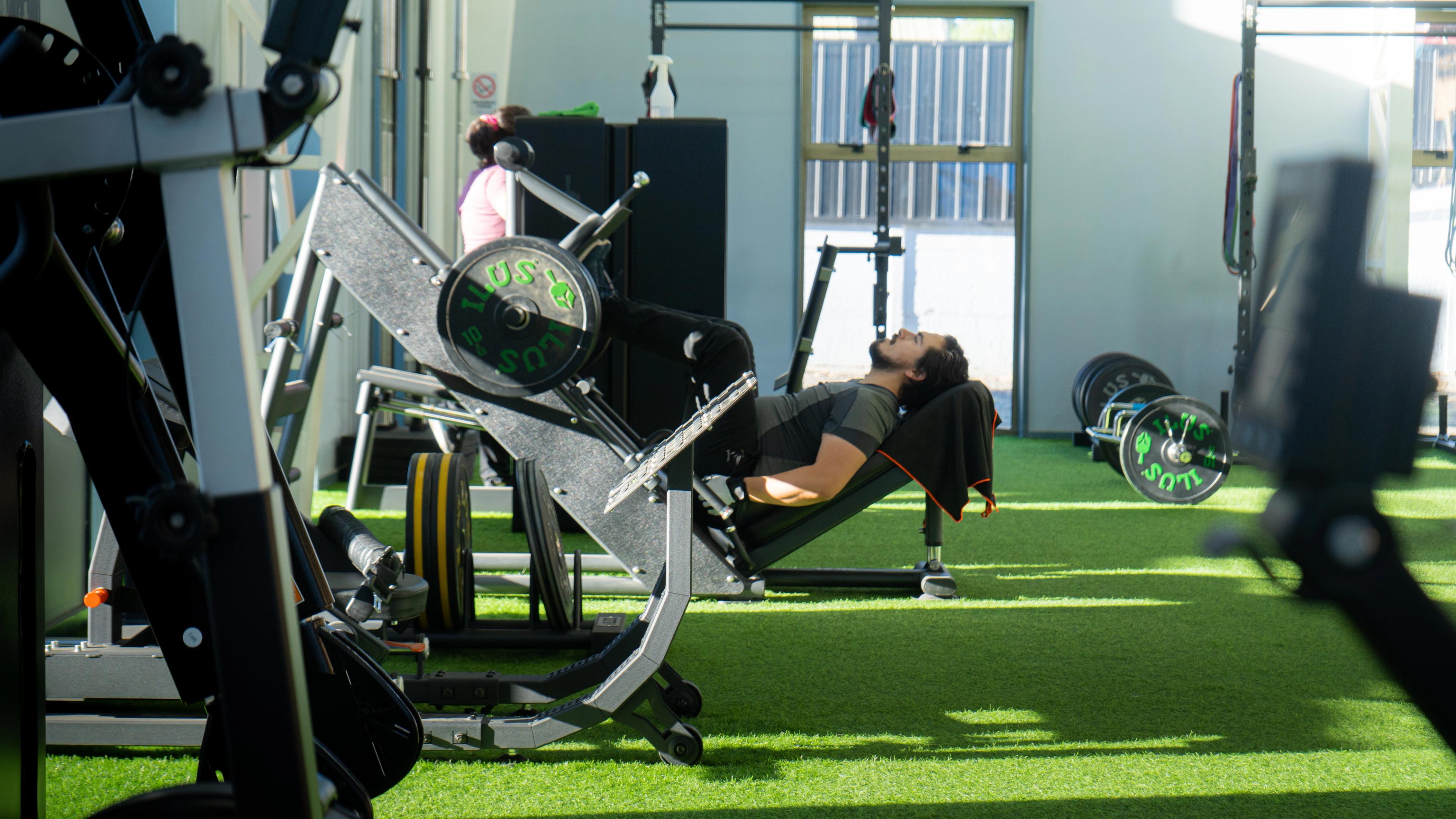 Una persona hace ejercicio en una máquina de prensa de piernas en un gimnasio bien iluminado con suelo de césped artificial.