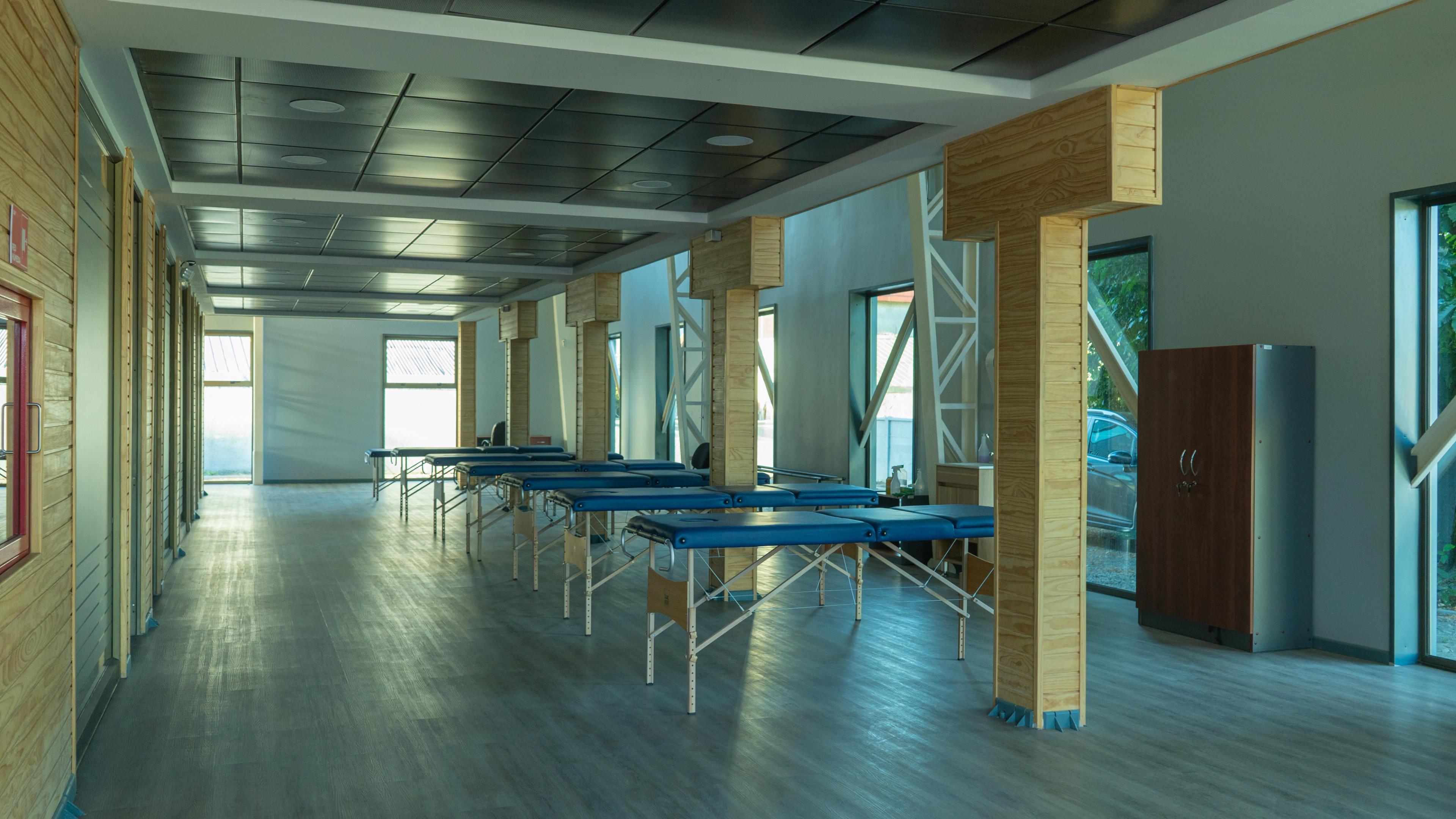 Una sala espaciosa con una fila de camillas de masaje azules vacías, pilares de madera y grandes ventanas que permiten la entrada de luz natural.