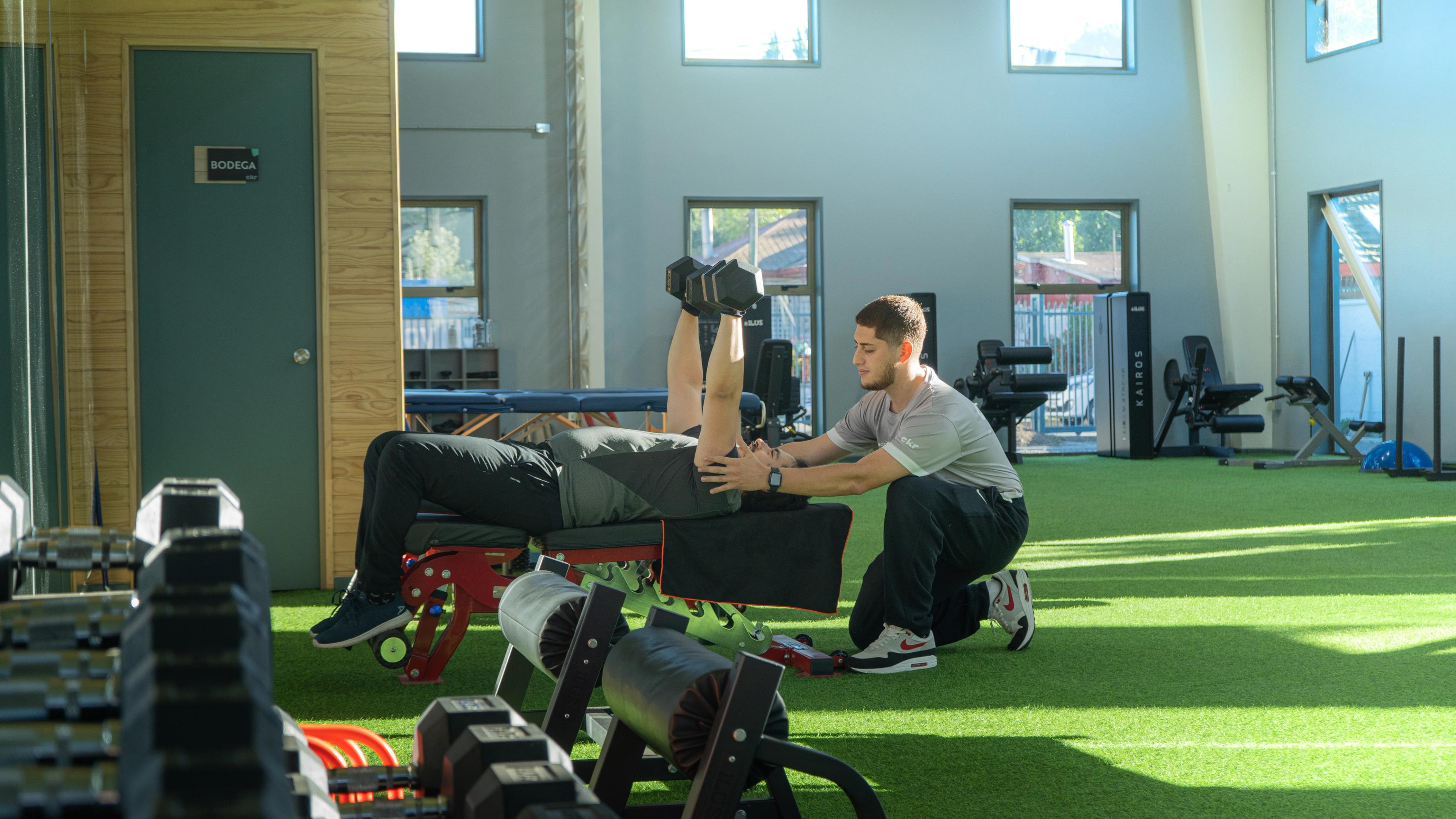 Un entrenador personal ayuda a un hombre a levantar mancuernas en un banco en un gimnasio luminoso con grandes ventanas y suelo verde.
