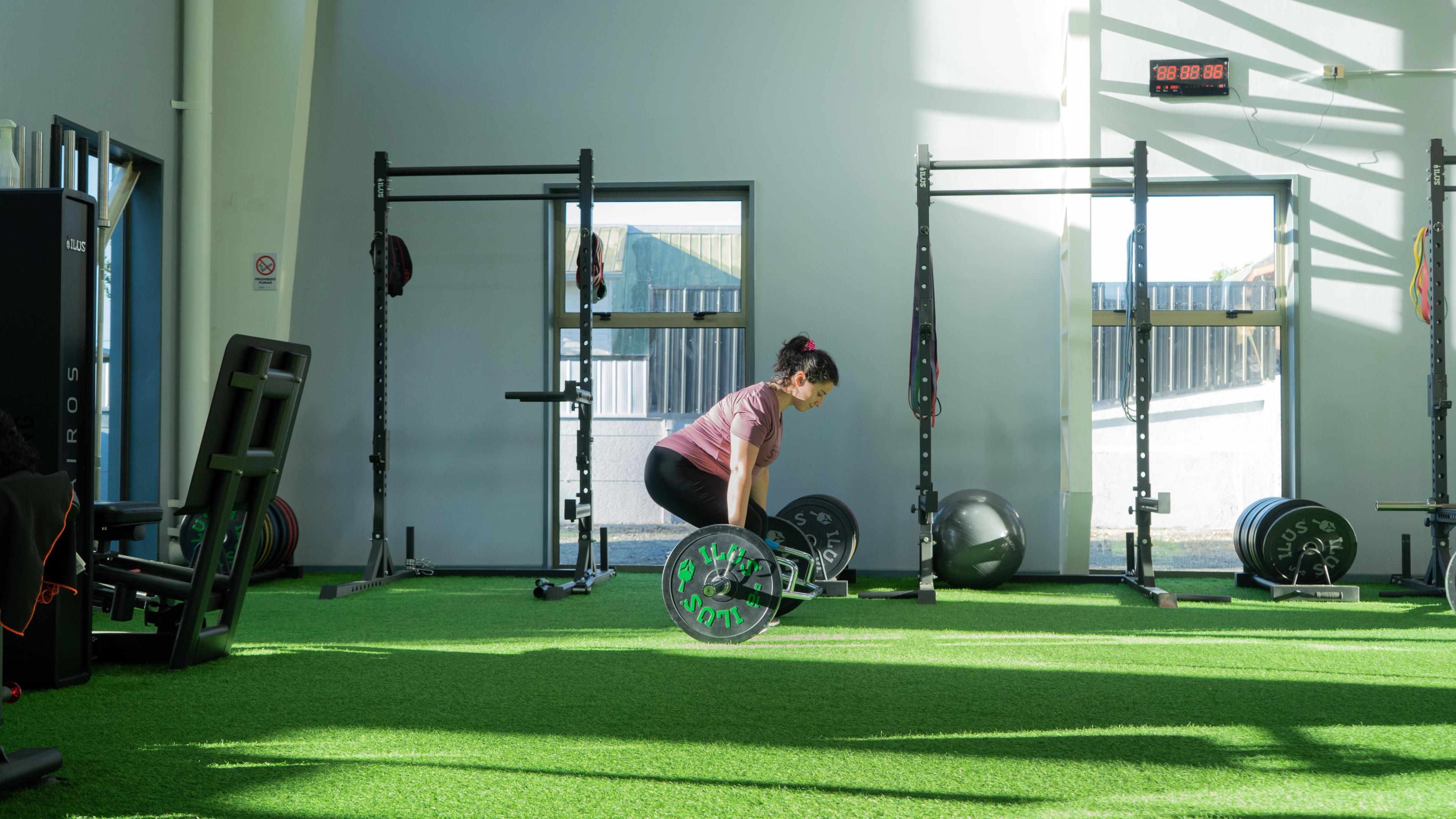 Una persona con ropa deportiva está haciendo peso muerto en un gimnasio soleado con suelo de césped artificial.
