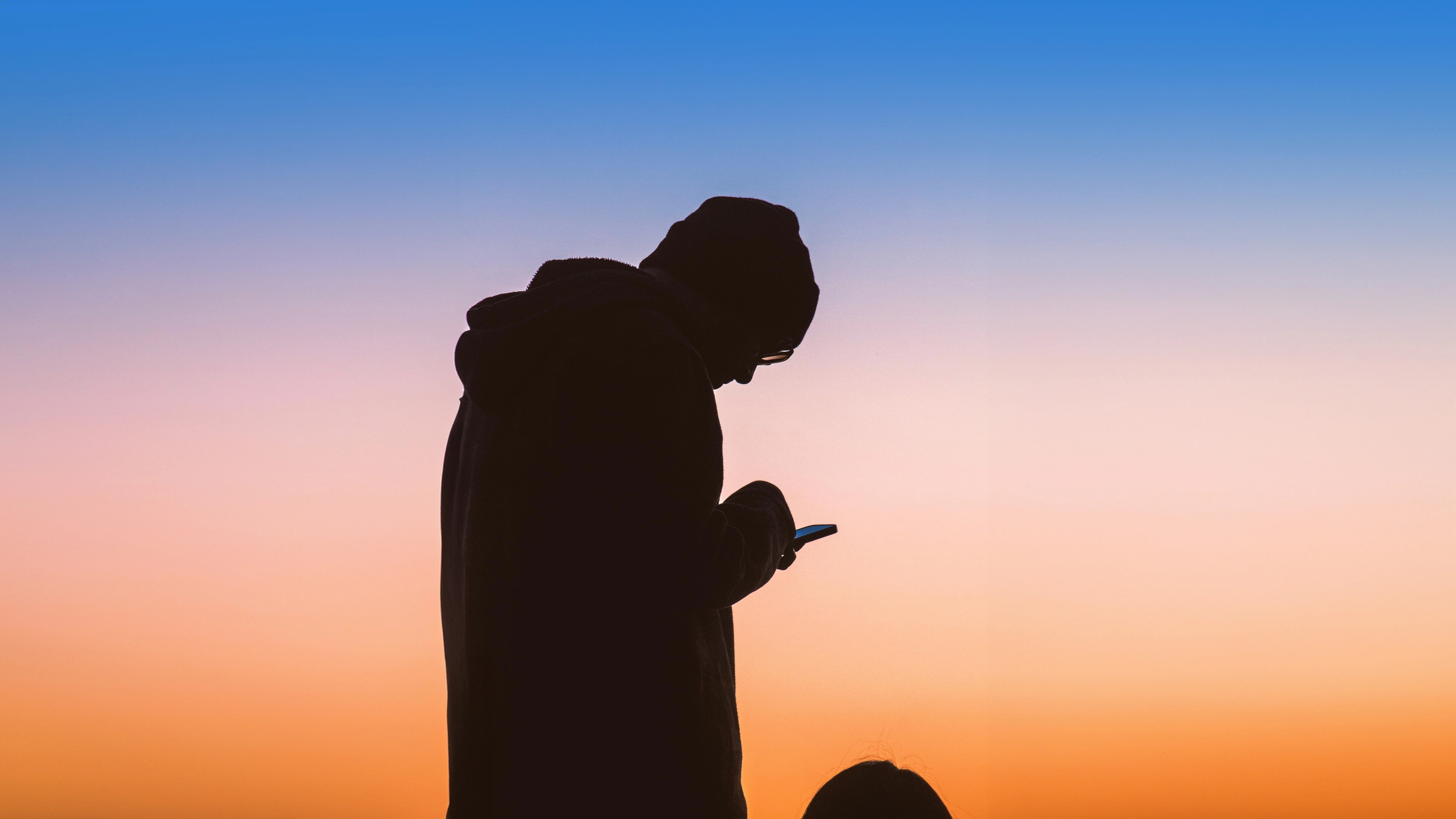 a shadowy figure looks at his phone with the sunset behind him.