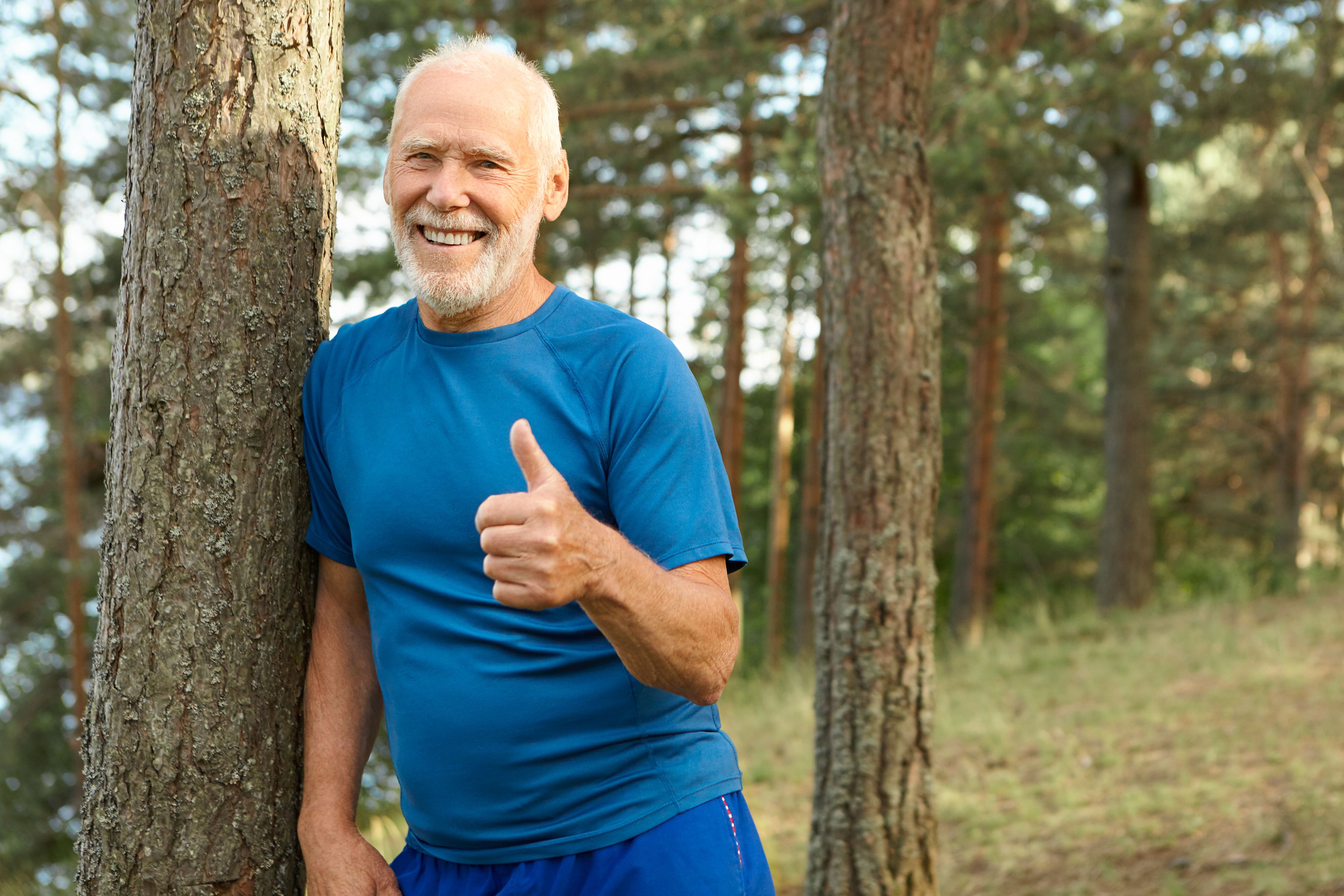 Active senior man smiling outdoors and giving a thumbs-up in a forest setting, representing healthy aging, vitality, fitness, and an active lifestyle