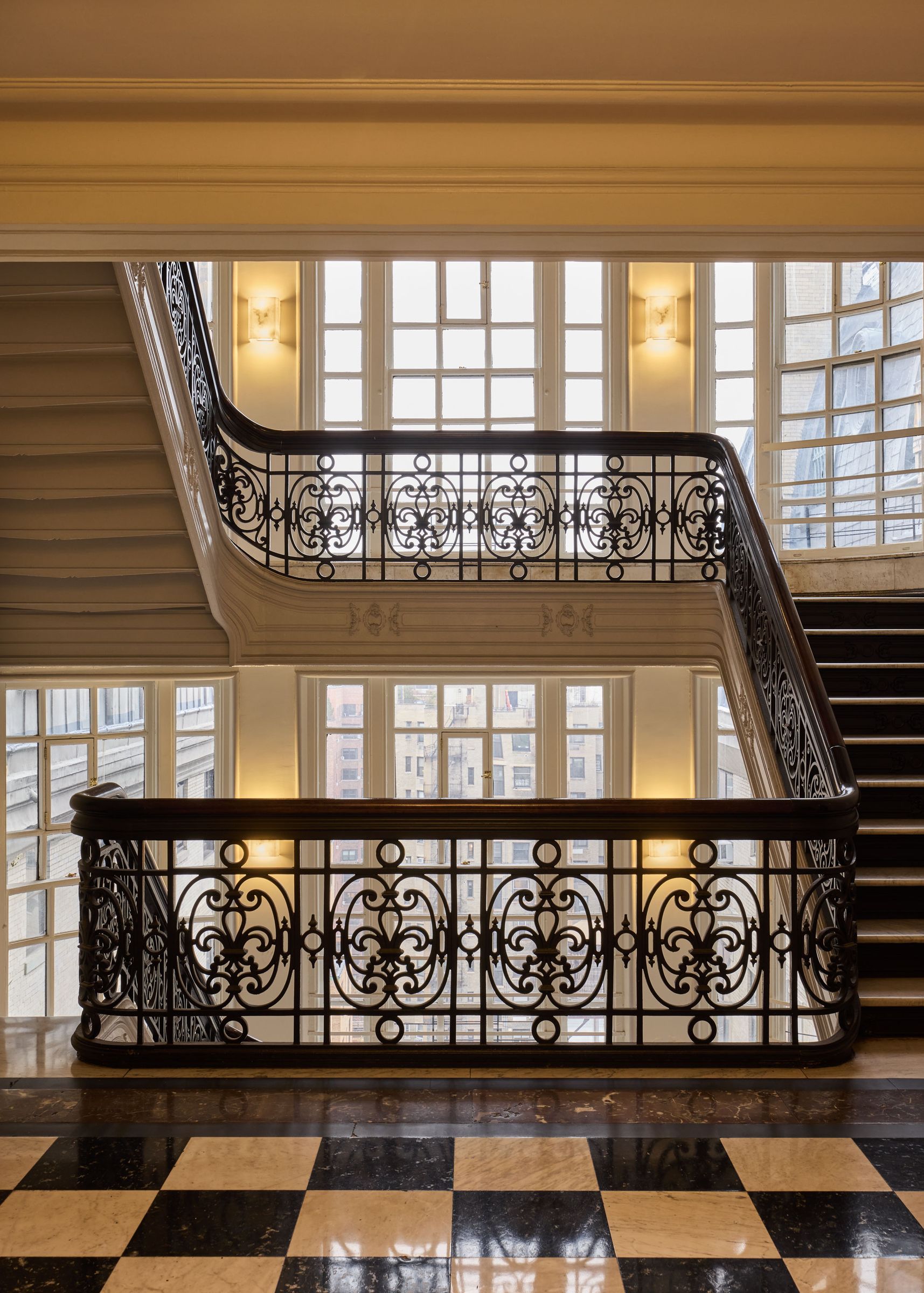 The landing of a grand staircase with an ornate black iron railing, set against a wall of windows on a checkerboard floor.