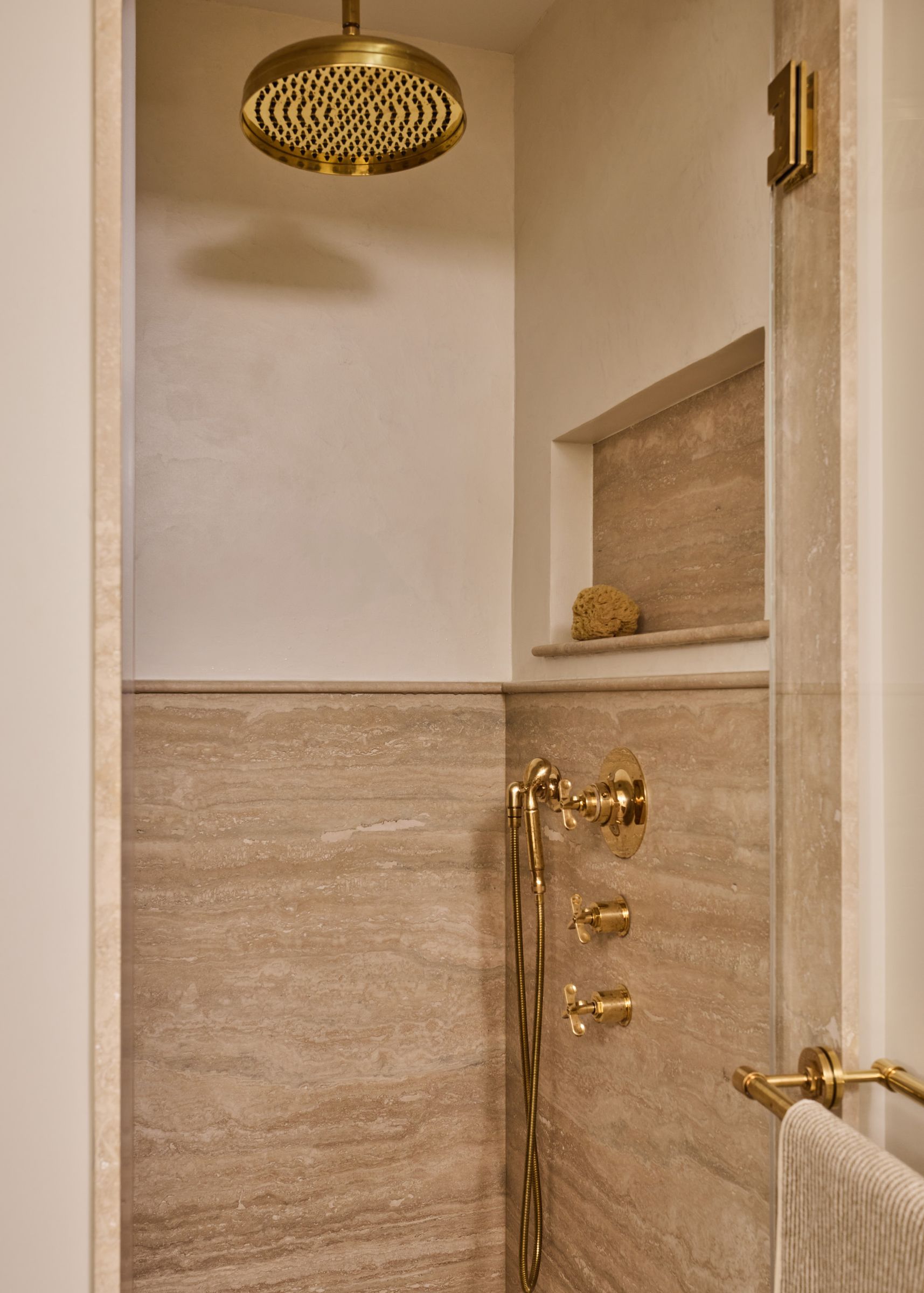 A close-up of a walk-in shower with beige stone walls and luxurious brass fixtures, including a large rain shower head.