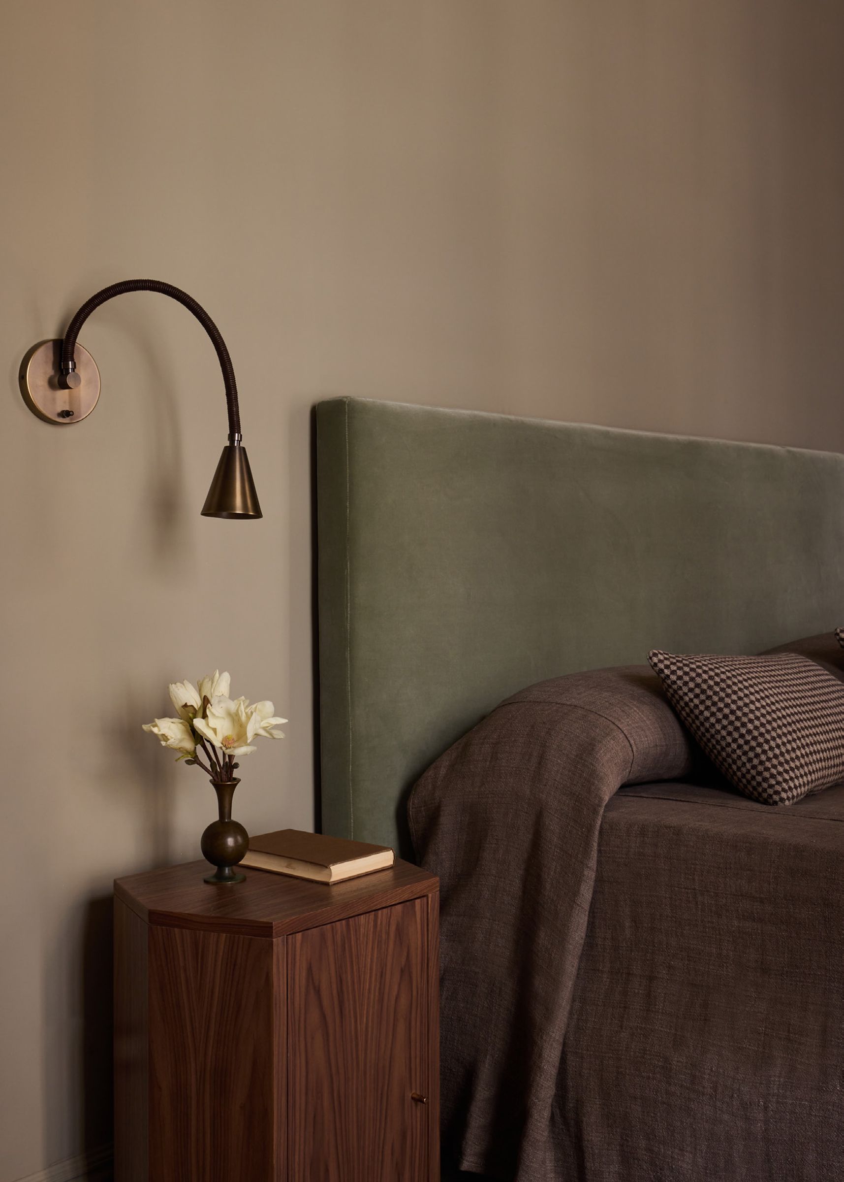 A close-up of a bedside table with a vase of flowers, next to a bed with a green velvet headboard and a wall lamp.