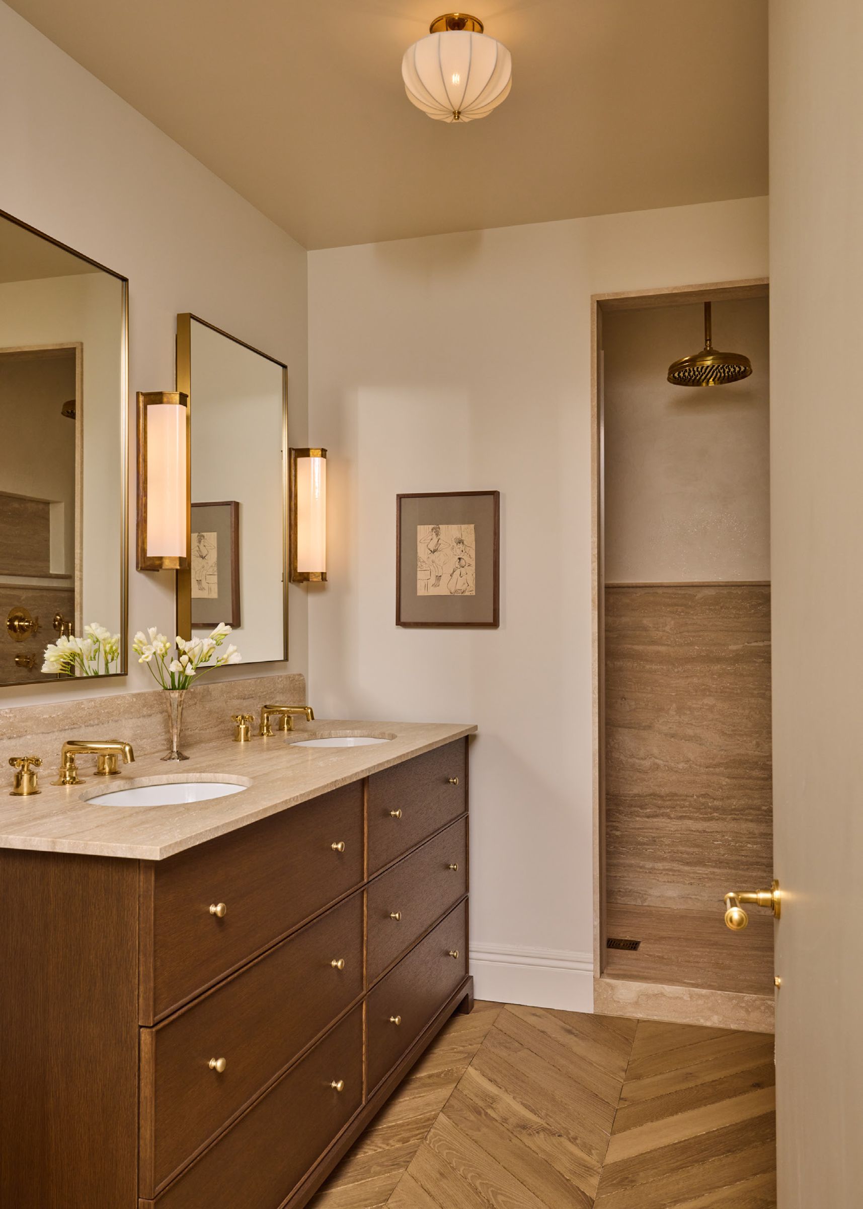 A bathroom with herringbone wood floors, a long wood double vanity with a stone top, and a view into a walk-in shower.