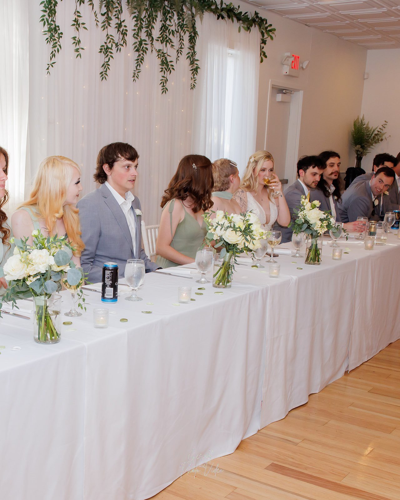 Long head table with white linens and low floral centerpieces at a wedding reception at The Arbor Event Venue