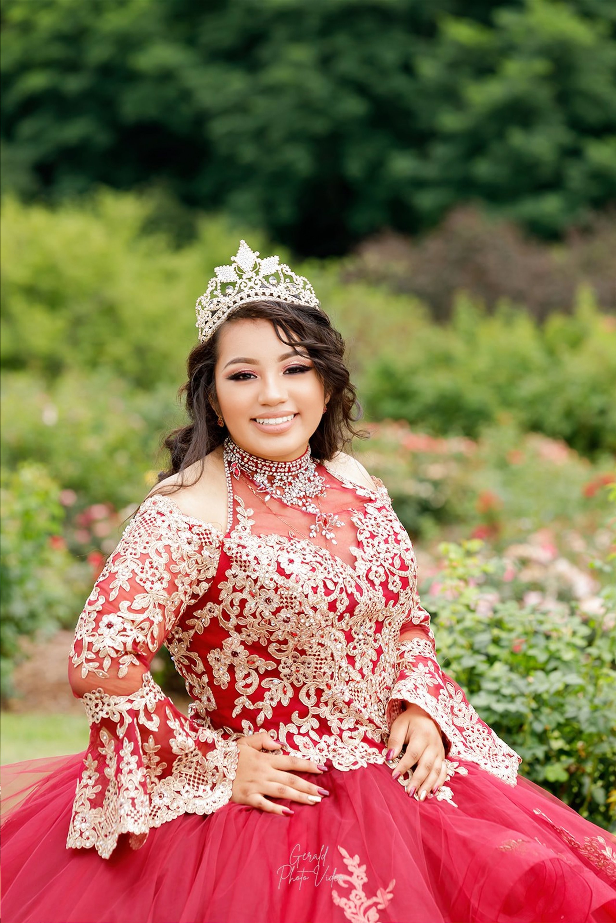 Quinceañera in a red gown with gold lace sleeves smiling in a garden, Omaha quinceañera portrait