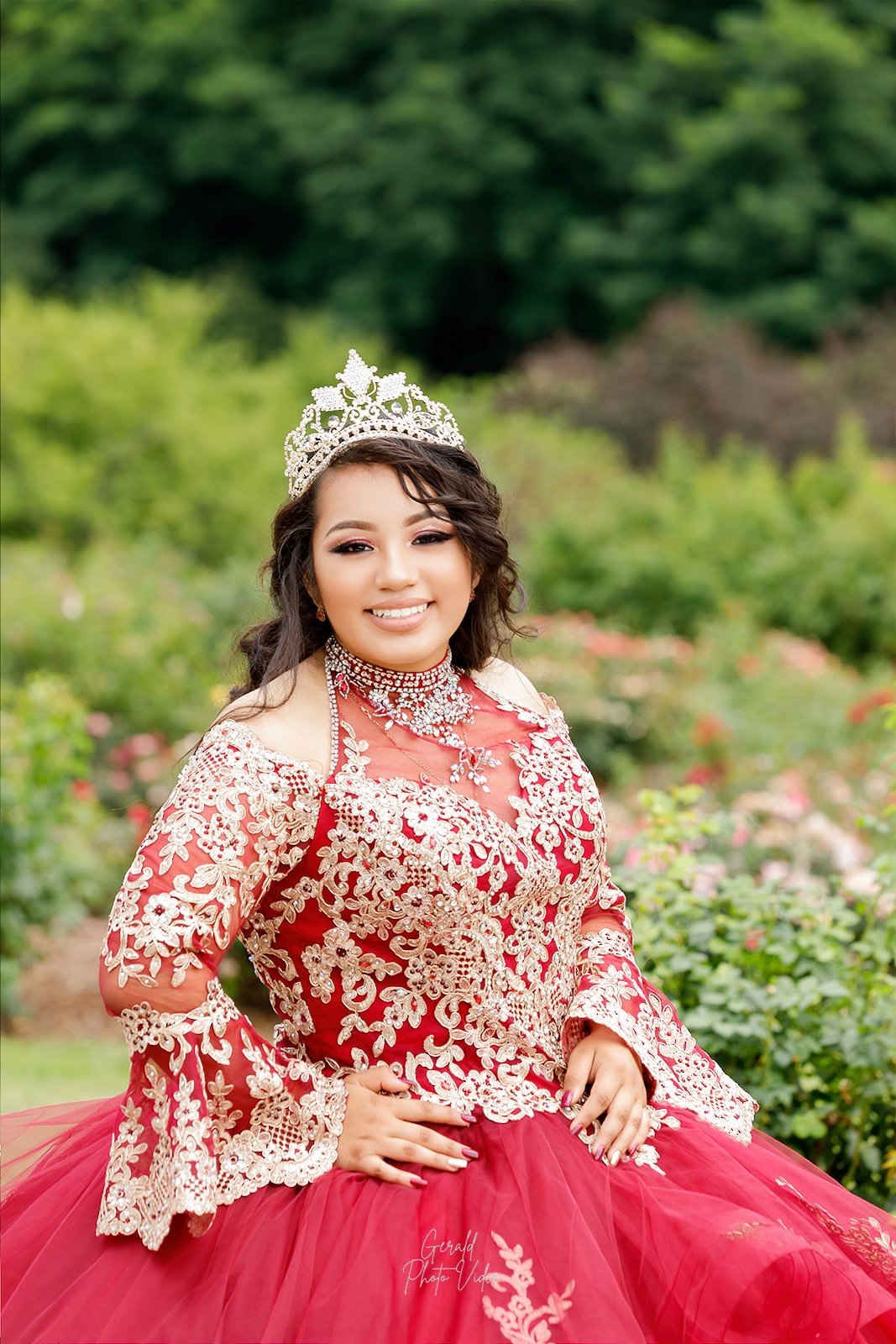 Quinceañera in a red gown with gold lace sleeves smiling in a garden, Omaha quinceañera portrait