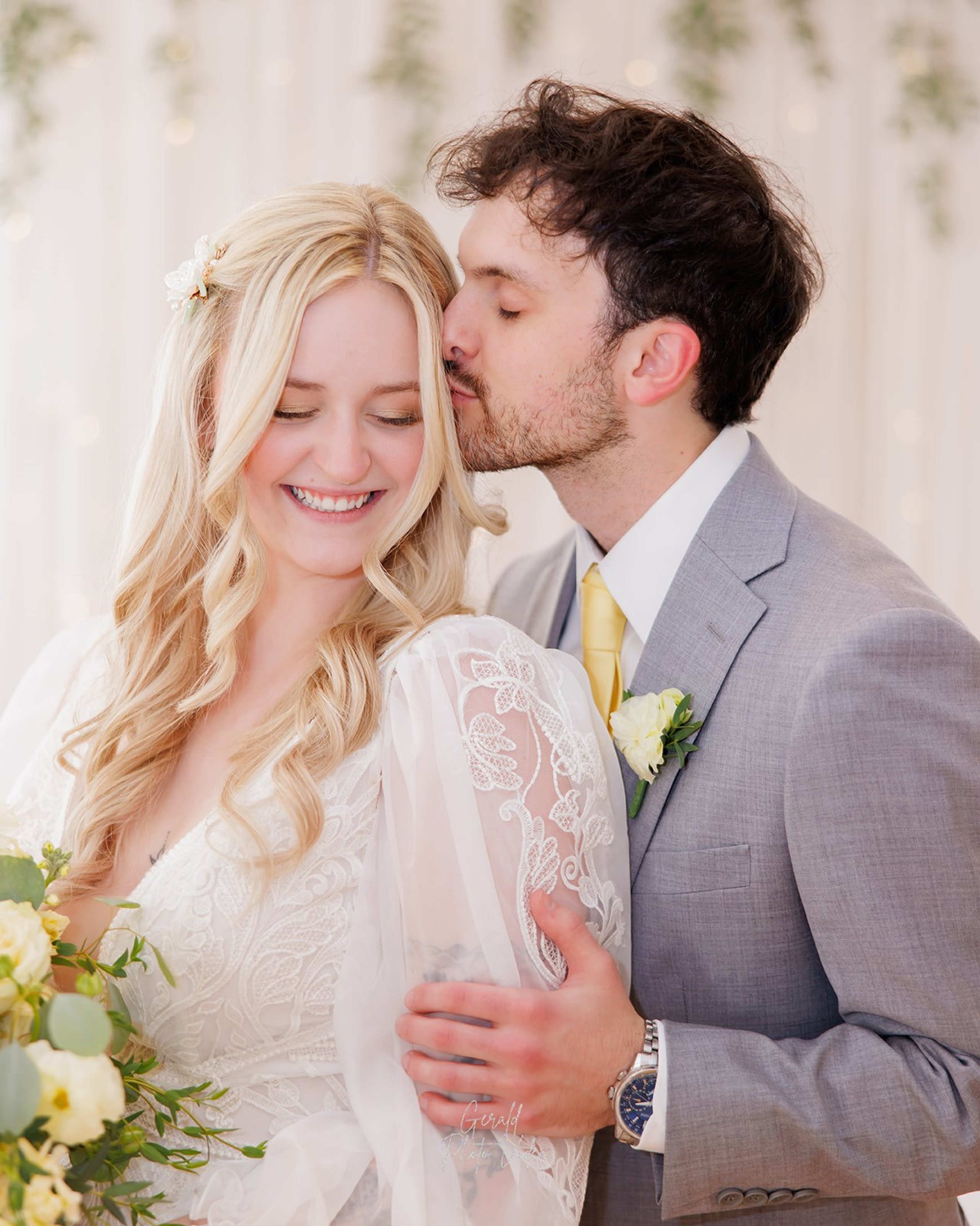 John kissing Logan on the cheek as she smiles in front of sheer drapes with hanging greenery