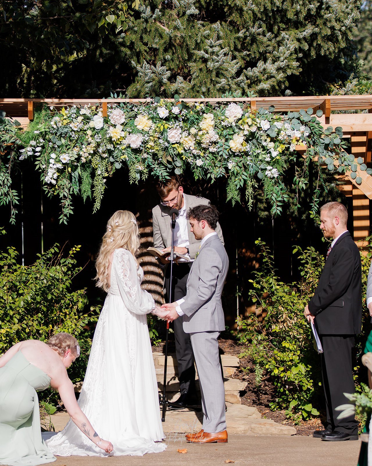 Logan and John holding hands at the altar under a floral arch with greenery at The Arbor Event Venue in Omaha