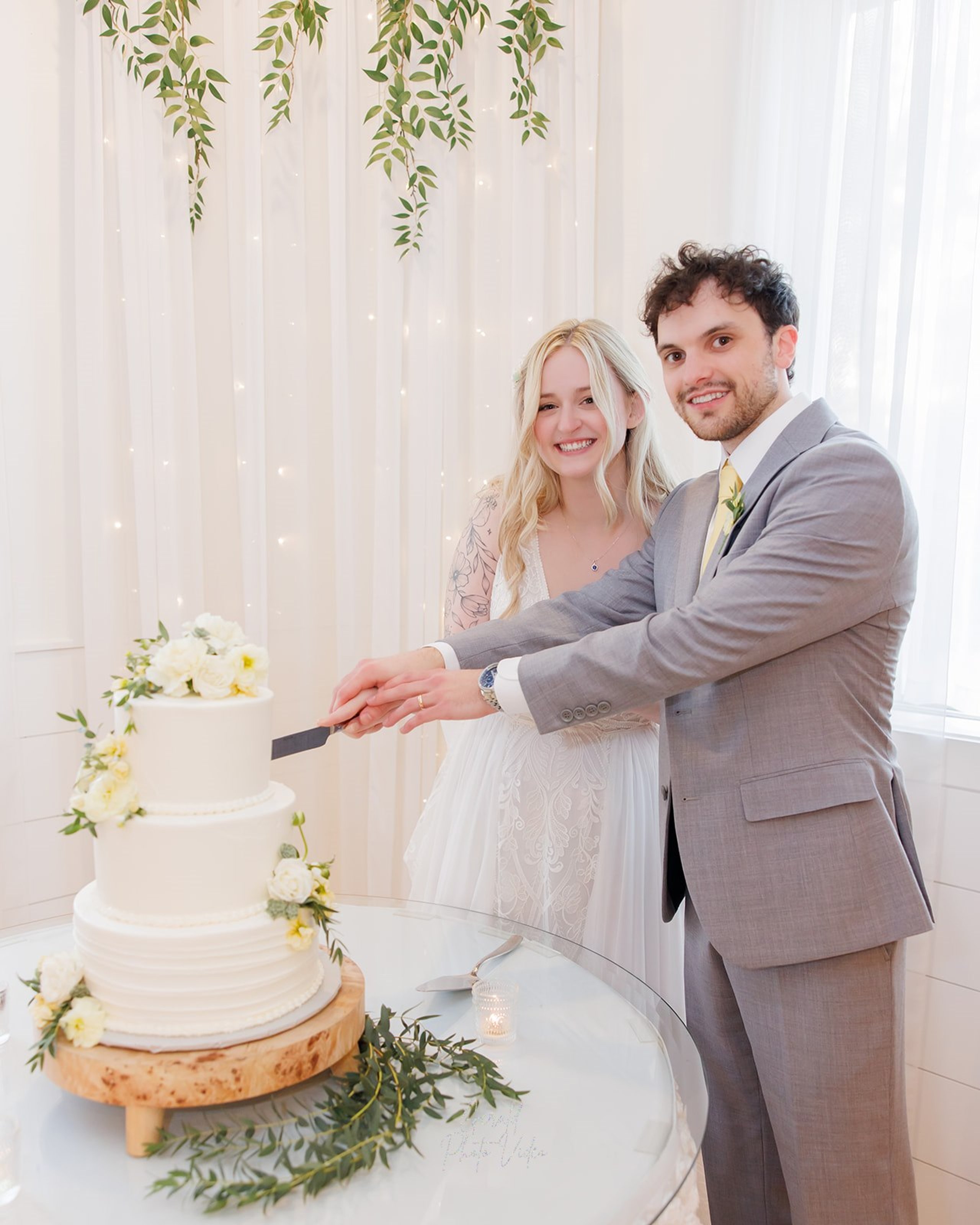 Logan and John cutting their white three tier wedding cake decorated with flowers and greenery