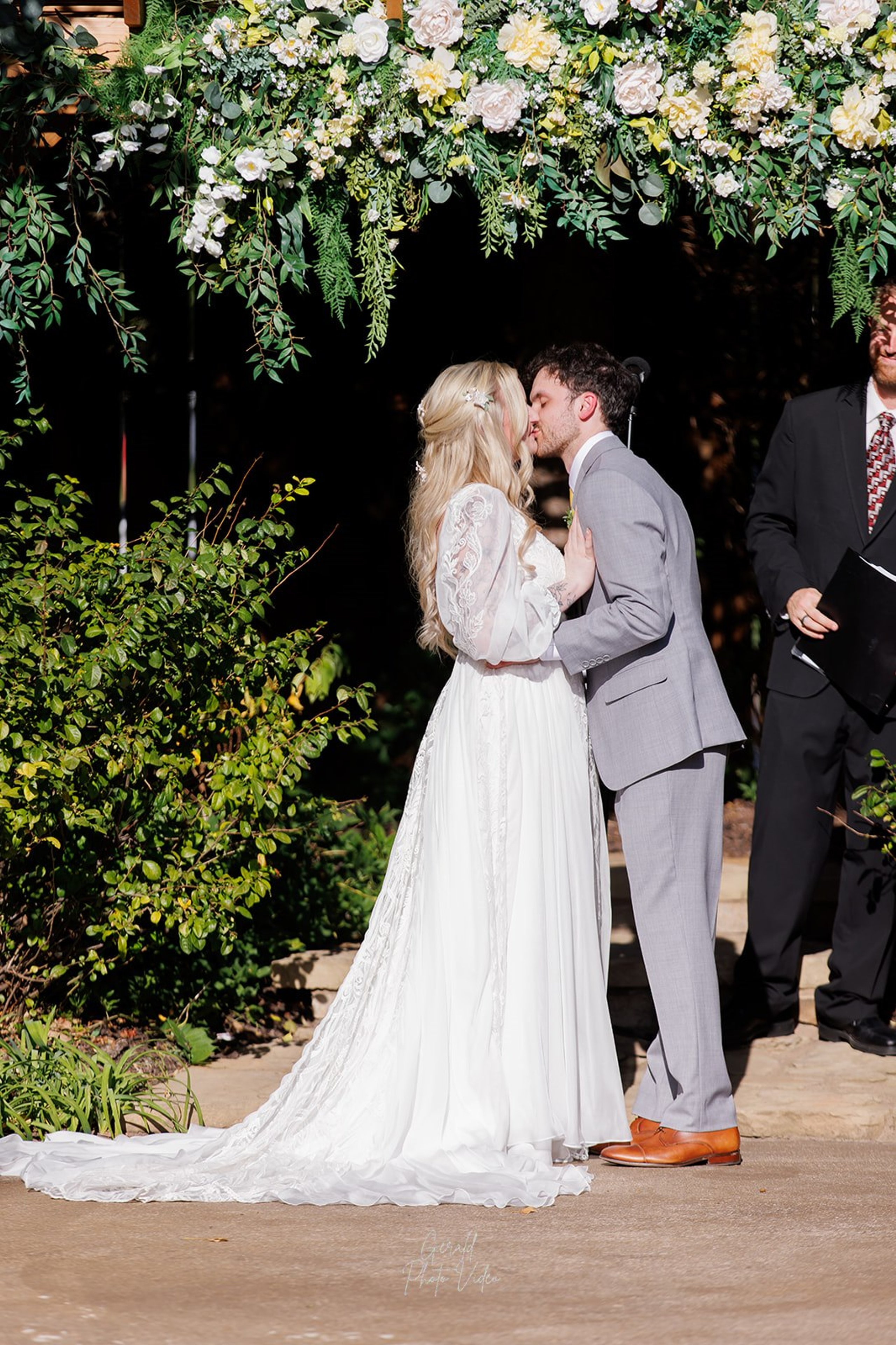 Logan and John sharing their first kiss as newlyweds under a white and green floral arch at The Arbor Event Venue, Omaha