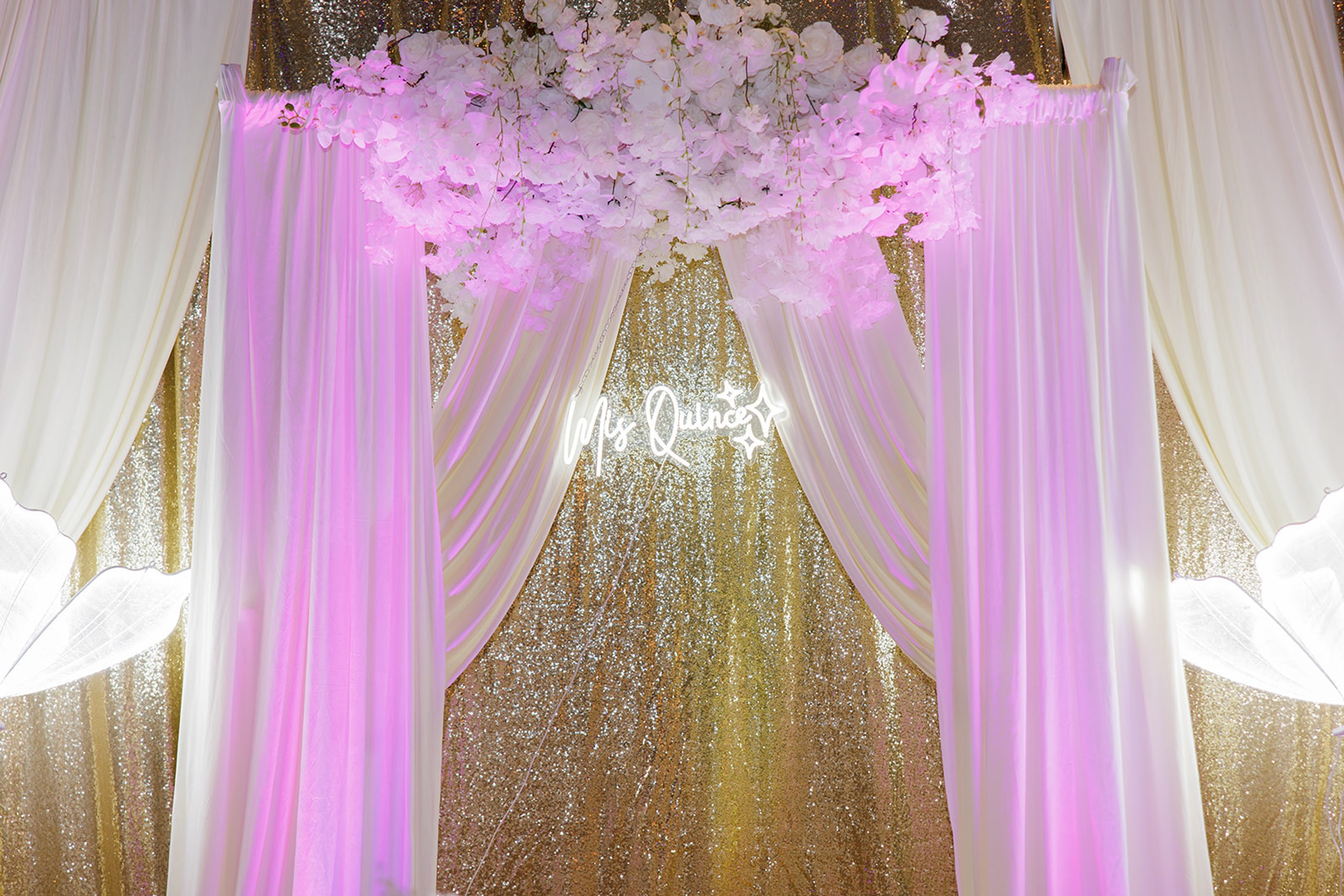 Arco floral con cortinas blancas, lentejuelas doradas y un letrero de neón en una quinceañera en Admiral Theater, Omaha